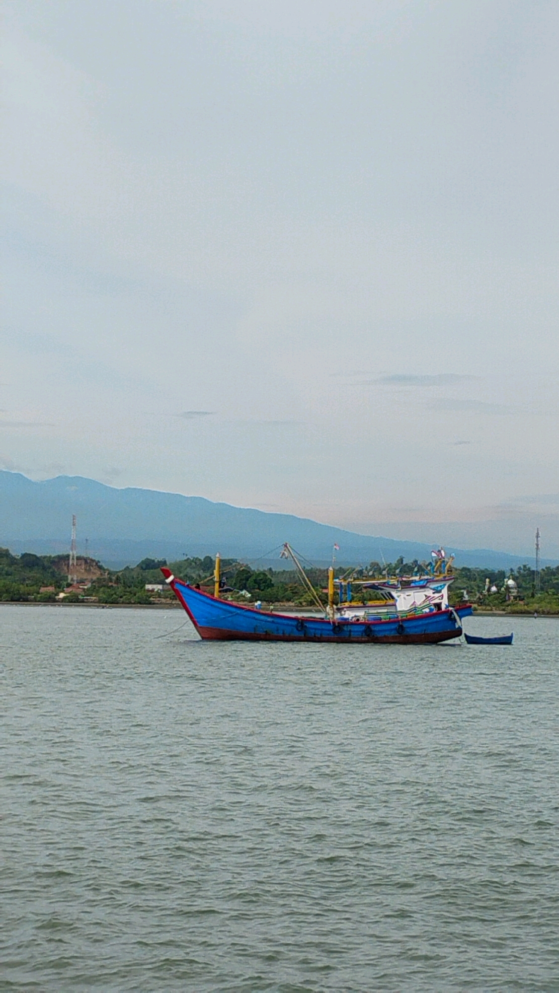 A blue and red fishing boat is anchored in calm, greenish-grey water. The boat is long and narrow, with a red bow and a blue hull. It has a white superstructure with various poles and antennas. In the background, a low, green, treed shoreline is visible, with some structures and what appears to be a tower. Further back, a range of hazy blue mountains stretches across the horizon under a pale, overcast sky. The scene is likely located in or near Lhokseumawe, Indonesia, given the presence of a fishing boat and a coastal landscape typical of the region. There are no people visible in the image.