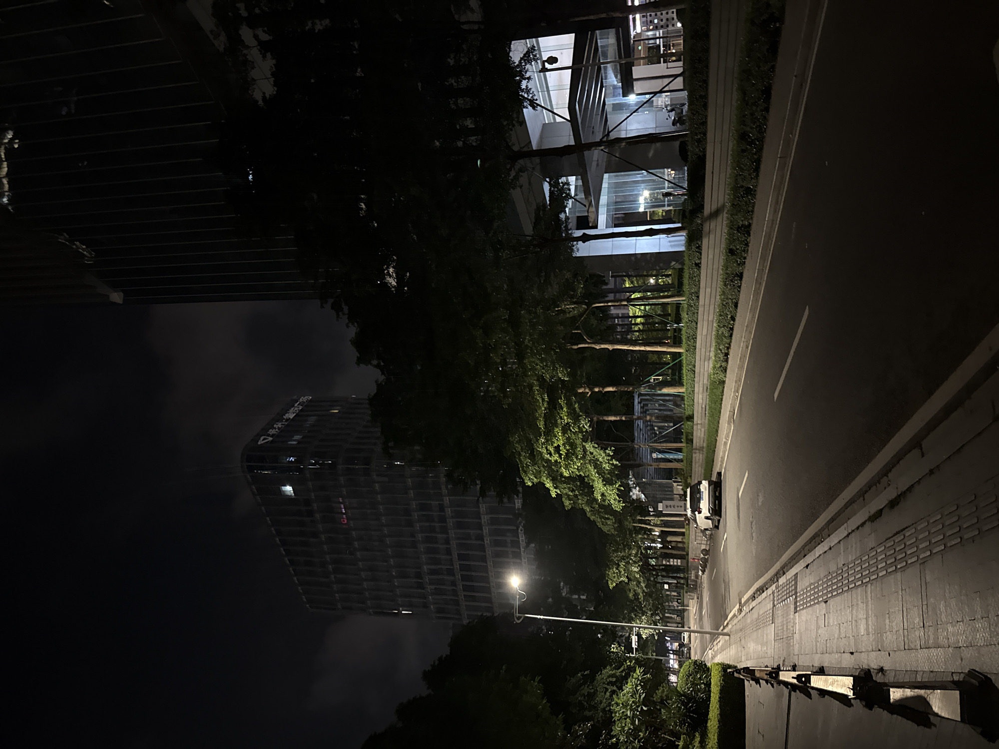 This media file depicts a nighttime street scene in Haizhu District, China. The foreground shows a paved sidewalk with tactile paving, bordering a dark asphalt road. The road is mostly empty, with a white vehicle parked on the left side. Lush green trees line the sidewalk, casting shadows and partially obscuring modern buildings.
To the left of the road, a multi-story building with a grid-like facade is visible, illuminated by interior lights. Further in the distance, a tall, dark skyscraper stands against a cloudy night sky. The skyscraper has a curved design and features illuminated windows and a visible logo or text, which appears to be in Chinese characters: "华科 影视广场".
A streetlamp illuminates a section of the sidewalk and the surrounding trees, creating pools of light and deep shadows, typical of a city at night. The overall atmosphere is quiet and serene, suggesting a peaceful evening in an urban environment. The presence of illuminated buildings and streetlights confirms that it is nighttime. There are no people clearly visible in the scene.