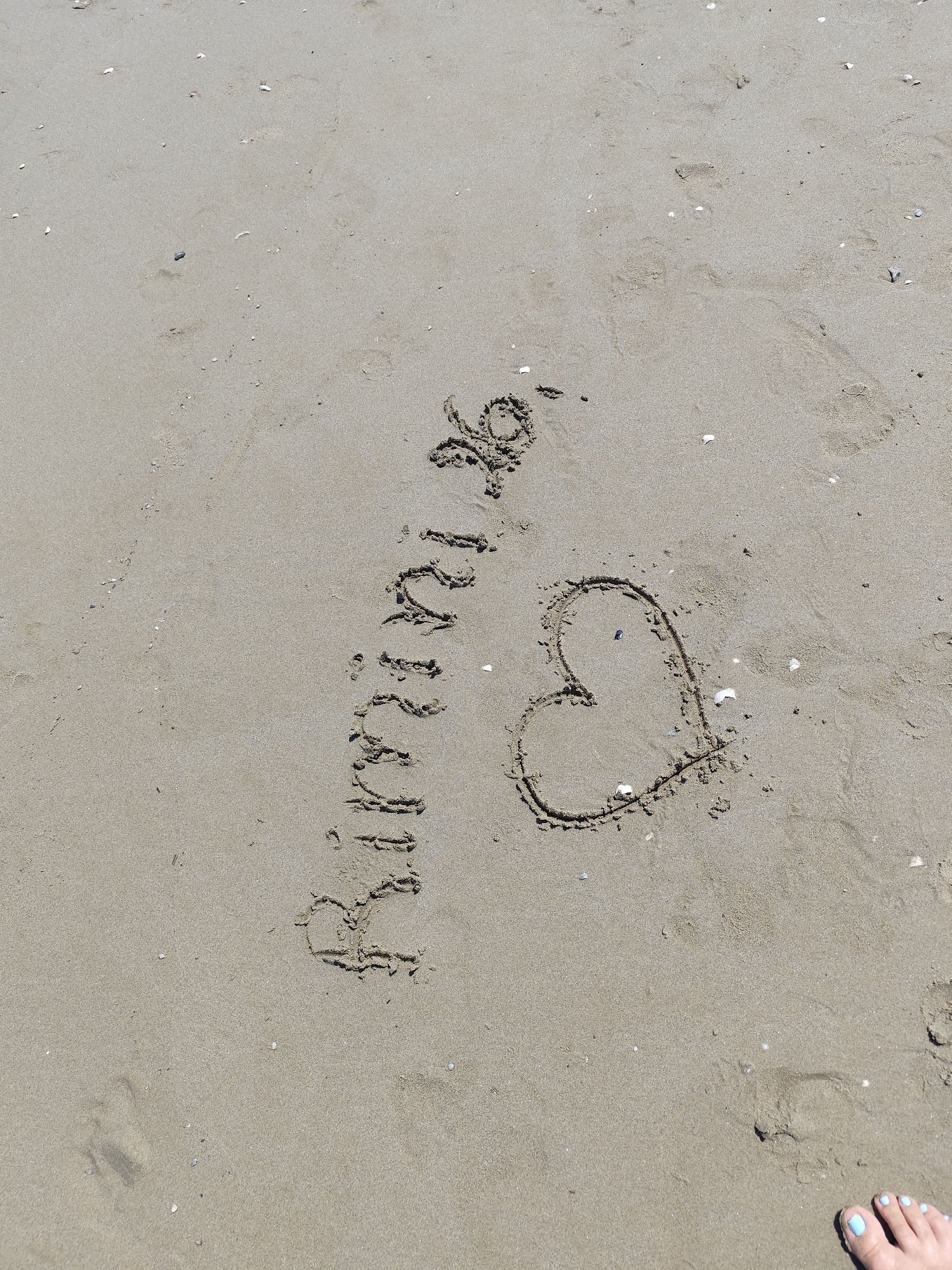 A close-up, overhead view of a sandy beach in Rimini, Italy, shows the word "Rimini" written in the sand, followed by a heart shape. The writing is done with a finger, creating a slightly uneven but clearly legible inscription. Small fragments of seashells are scattered across the sand. In the lower right corner, a foot with light blue painted toenails is partially visible, suggesting a person has just finished creating the message. The lighting indicates it is daytime, likely sunny.