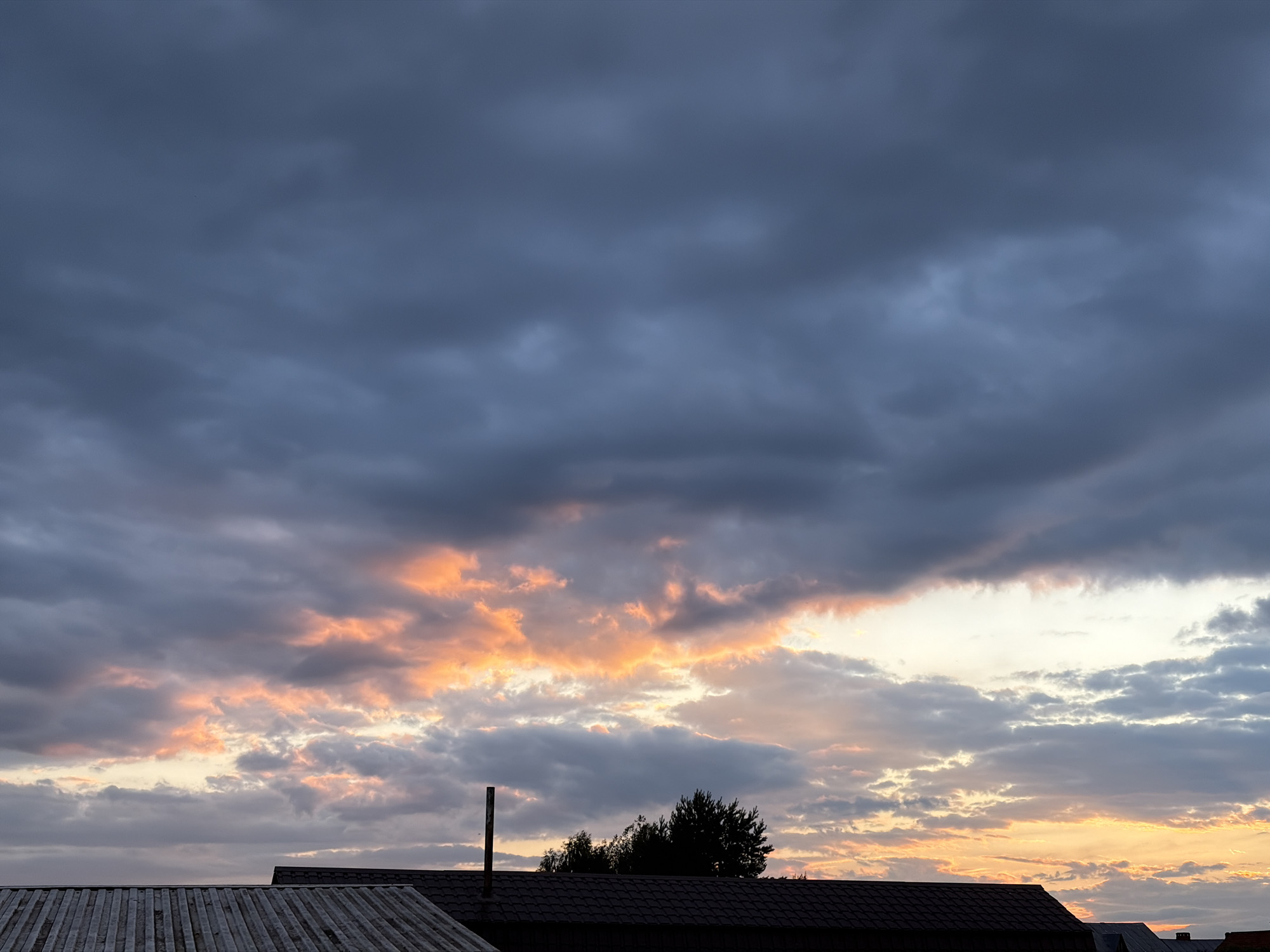 The image shows a cloudy sky at dusk over a residential area. The clouds are dark gray and blue, with patches of orange and pink light from the setting sun breaking through. Silhouetted against the sky are the roofs of several houses, some with visible chimneys. In the foreground, there is a corrugated metal roof. Trees are visible behind the houses. The scene is devoid of people or any visible activity.