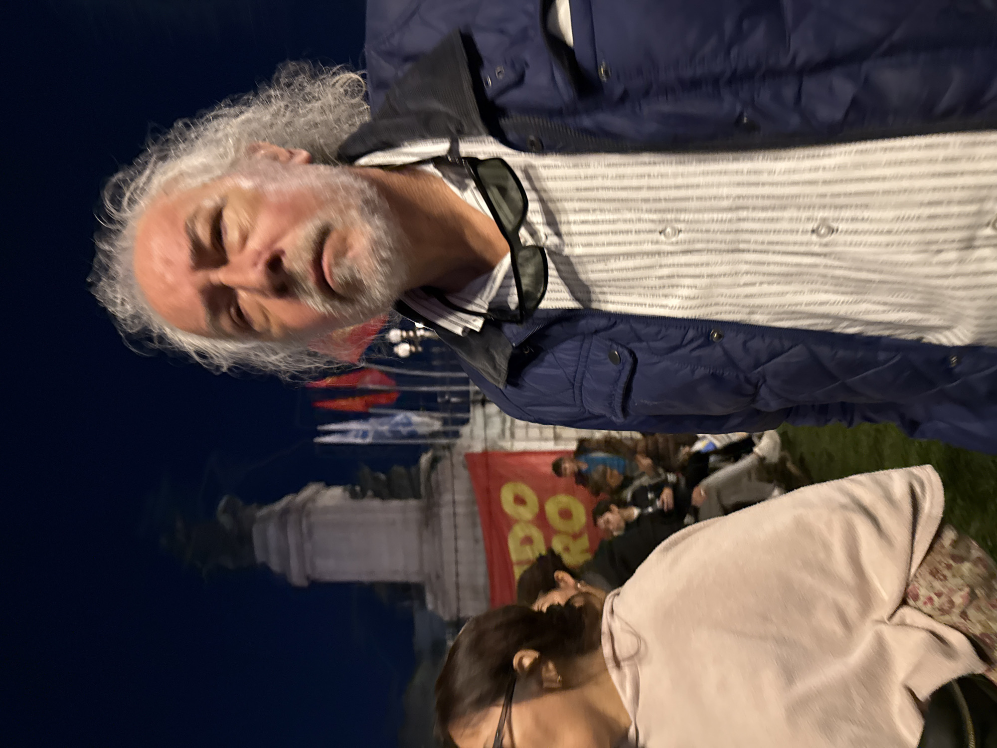 The image captures a scene outdoors at night in Buenos Aires, Argentina. In the foreground, a person with long gray hair and a beard is visible. They are wearing a blue jacket and a striped shirt with sunglasses tucked into the collar. A second person is also partially visible, wearing glasses and a light pink garment.
In the background, a group of people is gathered near a monument with flags. A red banner is visible with the words "DO DO RO" written on it in yellow. The monument itself is a light-colored stone structure. The overall environment is dark with some illumination from unseen light sources.