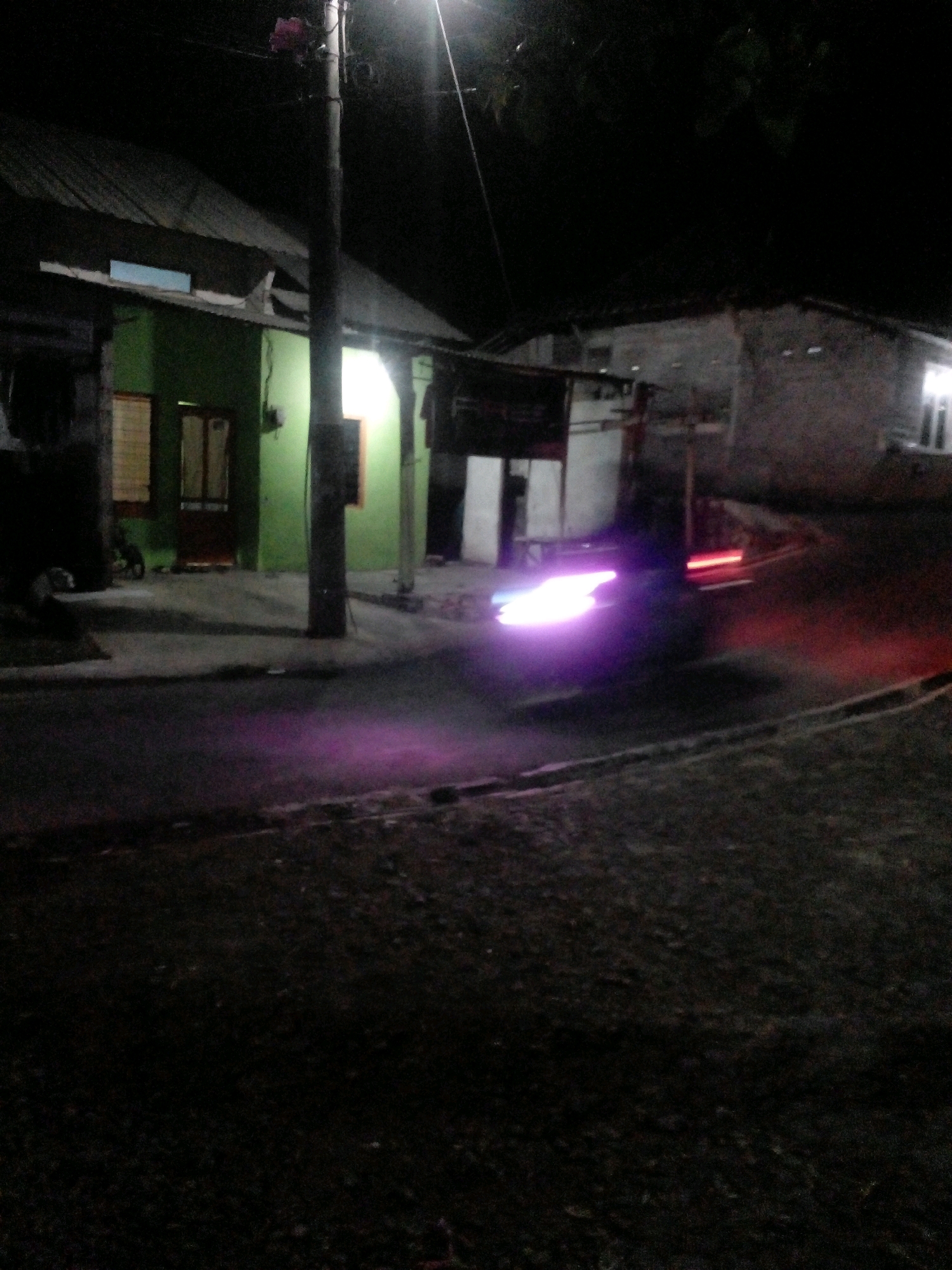 The image captures a street scene at night in Pungangan, Indonesia, dominated by artificial lighting and shadows. A prominent utility pole stands vertically on the left side of the frame, supporting electrical wires. Adjacent to the pole, a green building with a weathered, sloping roof is visible, featuring windows and a dark-colored door. To the right of this structure, another building with a lighter-colored, rough-textured wall is present, with illuminated windows suggesting occupancy. The street itself is paved and appears to be a moderately busy thoroughfare. A vehicle, though indistinct, is captured in motion, indicated by streaks of bright purple and red light trailing behind it, suggesting taillights and possibly exhaust fumes. The overall atmosphere is dimly lit, with the vehicle's lights providing the most significant illumination.
