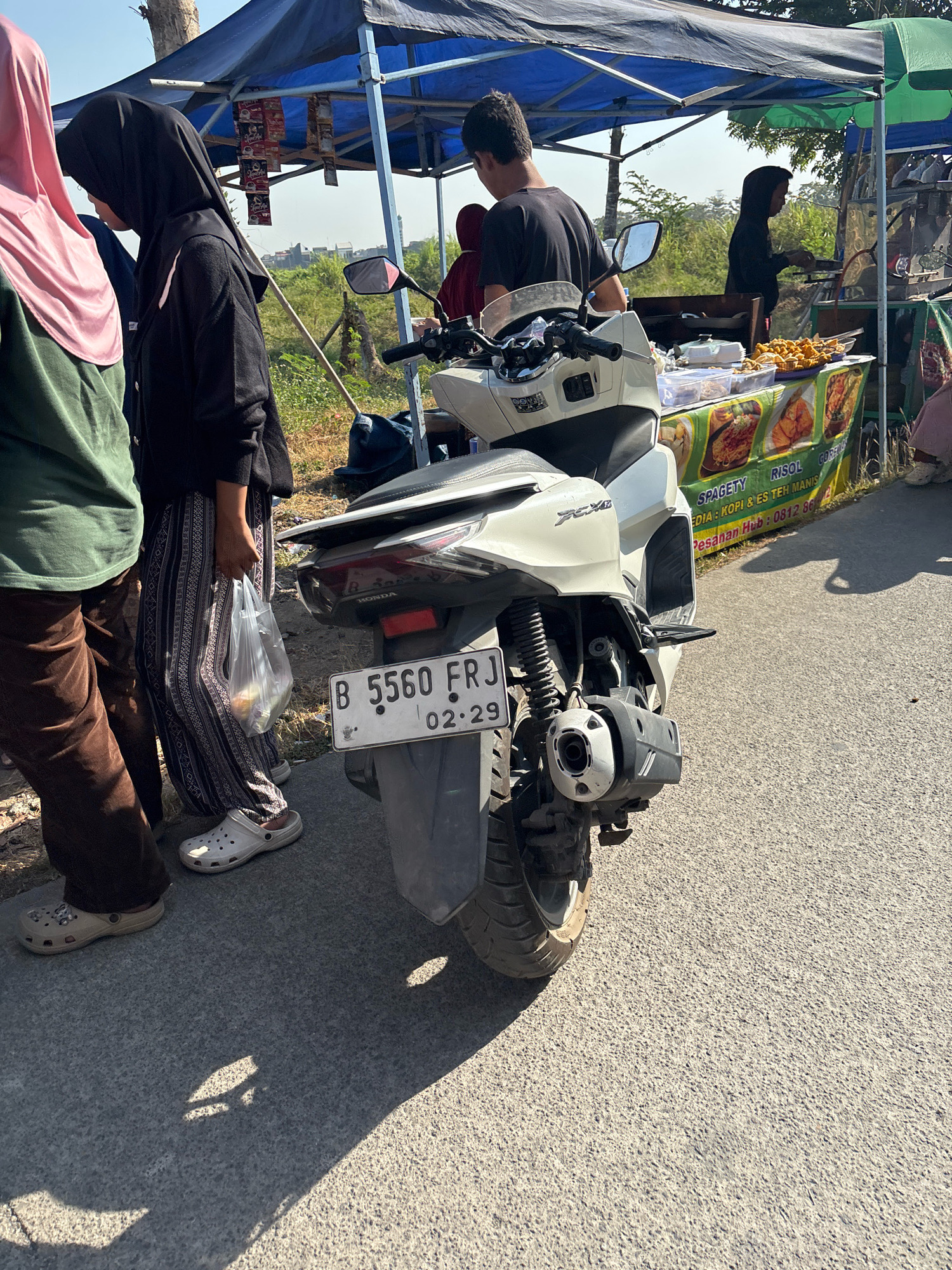 The image depicts an outdoor scene in Telagamurni, Indonesia, likely near a roadside stall. In the foreground, a white scooter is parked, with its license plate visible, reading "B 5560 FRJ" and "02-29". A person wearing dark patterned pants and white clogs stands next to the scooter, holding a clear plastic bag. To the left, another individual in a green shirt and brown pants is partially visible. Behind the scooter, a stall with a blue awning is set up, displaying various food items and drinks for sale. Several people are present around the stall, including one in a black hijab and another in a black t-shirt. The environment appears to be a sunny day with some vegetation and buildings visible in the background.