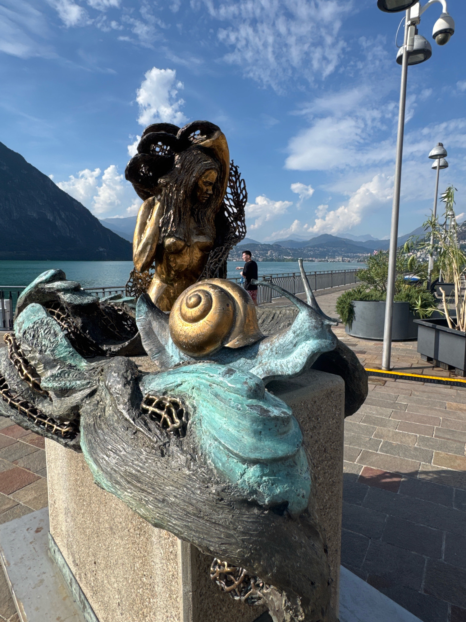 A bronze sculpture of a nude woman with flowing hair and a golden snail shell on her back is situated in the foreground of the image. The sculpture rests on a stone pedestal. The woman's body is partially obscured by a metallic net-like structure. The sculpture has a patina of green and bronze. In the middle ground, a man in a black shirt and red shorts stands by a metal railing overlooking a body of water. Beyond the water, a city with buildings and mountains can be seen in the distance under a partly cloudy blue sky. In the right side of the image, there are lampposts and potted plants. The overall scene is outdoors, suggesting a public space, likely a promenade or park area in Campione d'Italia, Italy, given the characteristic lakeside setting with mountains.