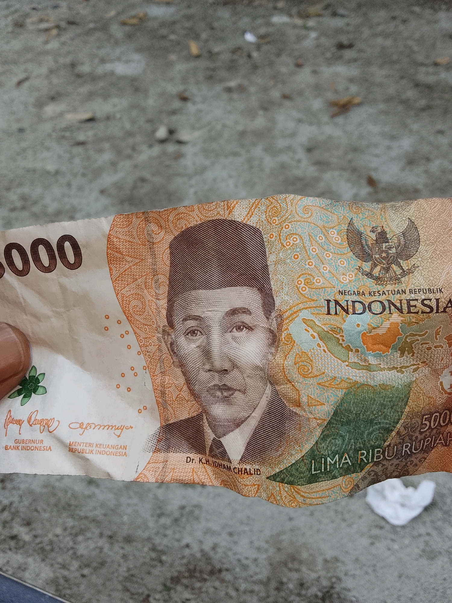A close-up, eye-level shot shows a hand holding a crumpled Indonesian 5,000 rupiah banknote. The banknote, featuring a portrait of a man in a hat and the Indonesian coat of arms, is predominantly orange, white, and teal. Text on the bill includes "5000", "NEGRA KESATUAN REPUBLIK", "INDONESIA", and "LIMA RIBU RUPIAH". The background is a blurred, greyish-brown textured surface, possibly concrete or asphalt, with some small, light-colored debris scattered across it. The lighting is even, suggesting an outdoor setting under diffuse light. No specific location in Padang Dalom, Indonesia can be determined from the image.