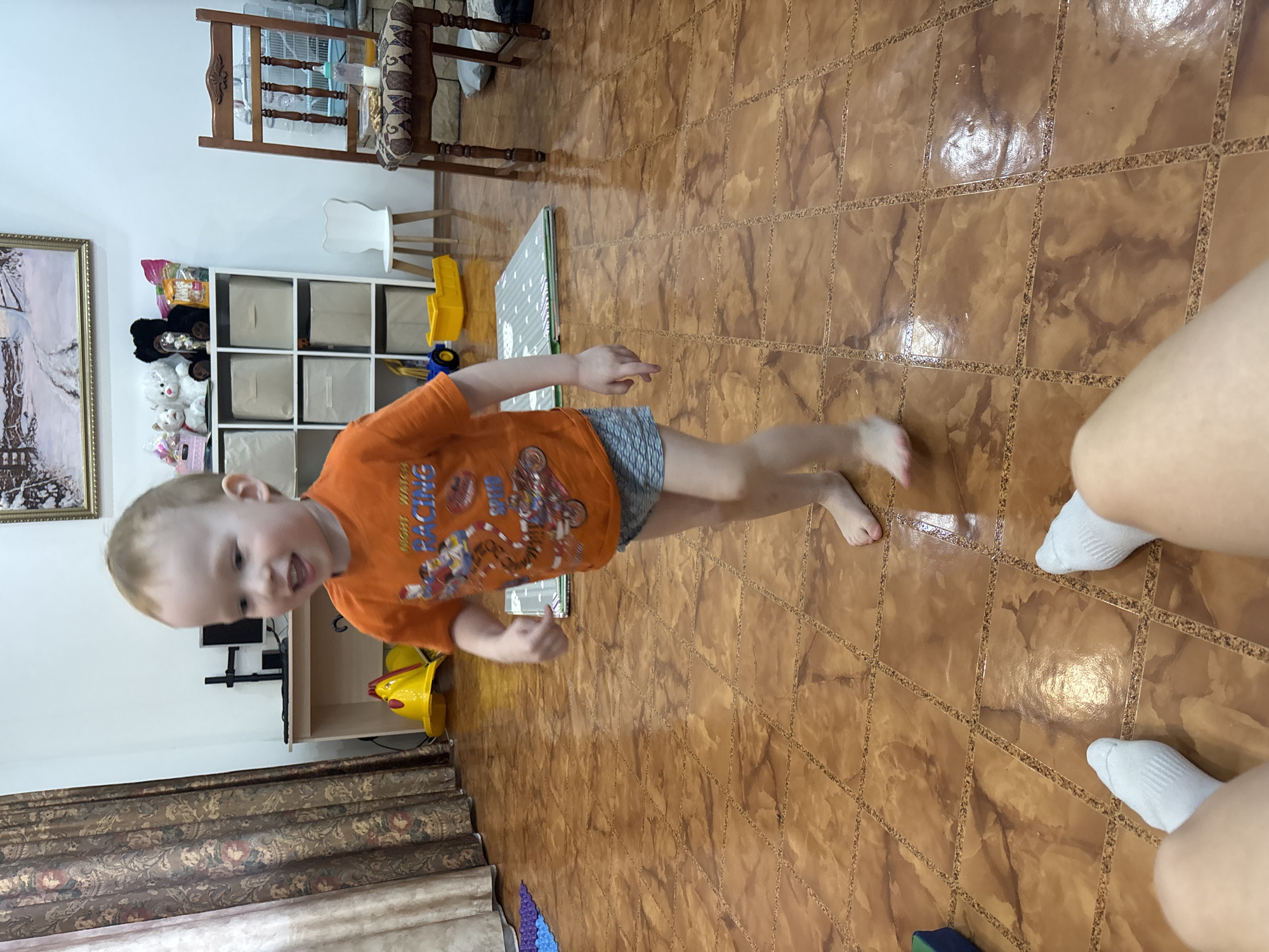 A young child in an orange shirt and shorts is running on a brown tiled floor. Near the child are legs of a person with white socks. In the background are furniture, toys, and a curtain. The location is implied to be in a home in Ферма, Russia.