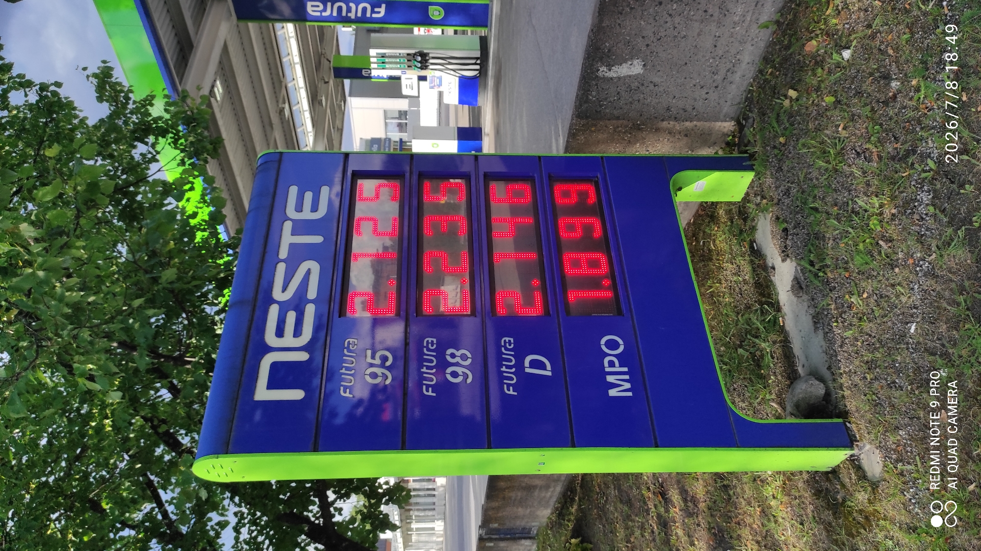 This is a vertical, eye-level shot of a Neste gas station price display in Iisalmi, Finland. The display features four columns showing fuel prices. From left to right, the prices are: 1.729 for Futura 95, 1.729 for Futura 98, 1.619 for Futura D, and 1.669 for MPO. The "NESTE" logo is prominently displayed on the left side of the sign. Behind the price display, the canopy of the gas station is visible with green and blue elements, along with gas pumps. The scene is outdoors, with a tree on the left providing shade and greenery. The ground is a mix of pavement, gravel, and grass. The timestamp in the lower right corner indicates the photo was taken on July 8, 2026, at 18:49. The lighting suggests it is late afternoon or early evening. There are no people visible in the frame. The sky is overcast. The text "FUTURA" is visible at the top of the gas station canopy in Finnish.