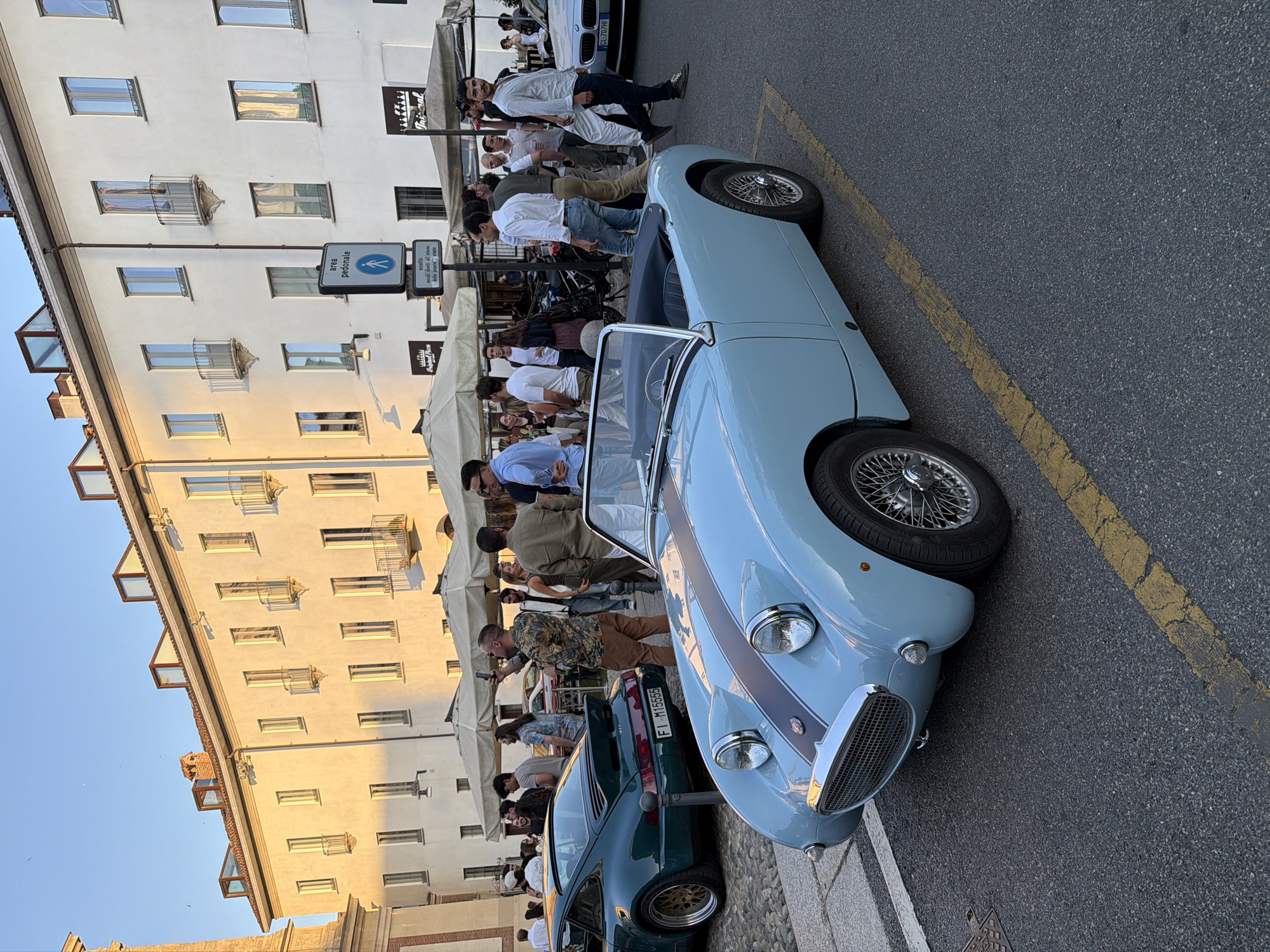 A light blue convertible classic car is parked on an asphalt road with yellow lane markings. The car has chrome wire wheels and a dark stripe on its hood. Behind the car, a group of people are gathered, some standing and talking, others walking. Several buildings with cream-colored facades and windows are visible in the background. Some of the buildings have awnings extended over outdoor seating areas. A dark green sports car is parked to the left of the classic car. The scene appears to be a street event or gathering in a European city during the daytime, with the setting sun casting a warm glow on the buildings.