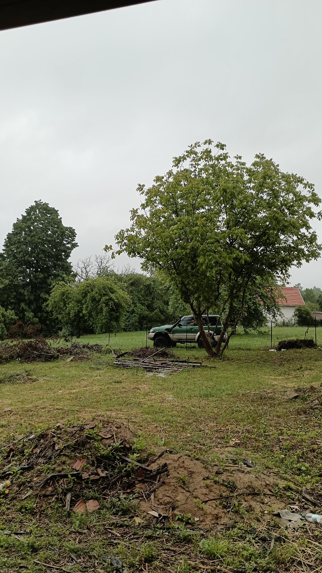 The image shows a rural scene, possibly in or near Mihaylovtsi, Bulgaria. Overcast sky. A green, off-road vehicle is parked partially obscured by a tree near a wire fence. In the background, there are other trees and a glimpse of a building with a red tile roof. The foreground features a grassy field with scattered debris, including piles of dirt, branches, and possible construction materials. No people are visible. The overall impression is one of untidiness or a work in progress.