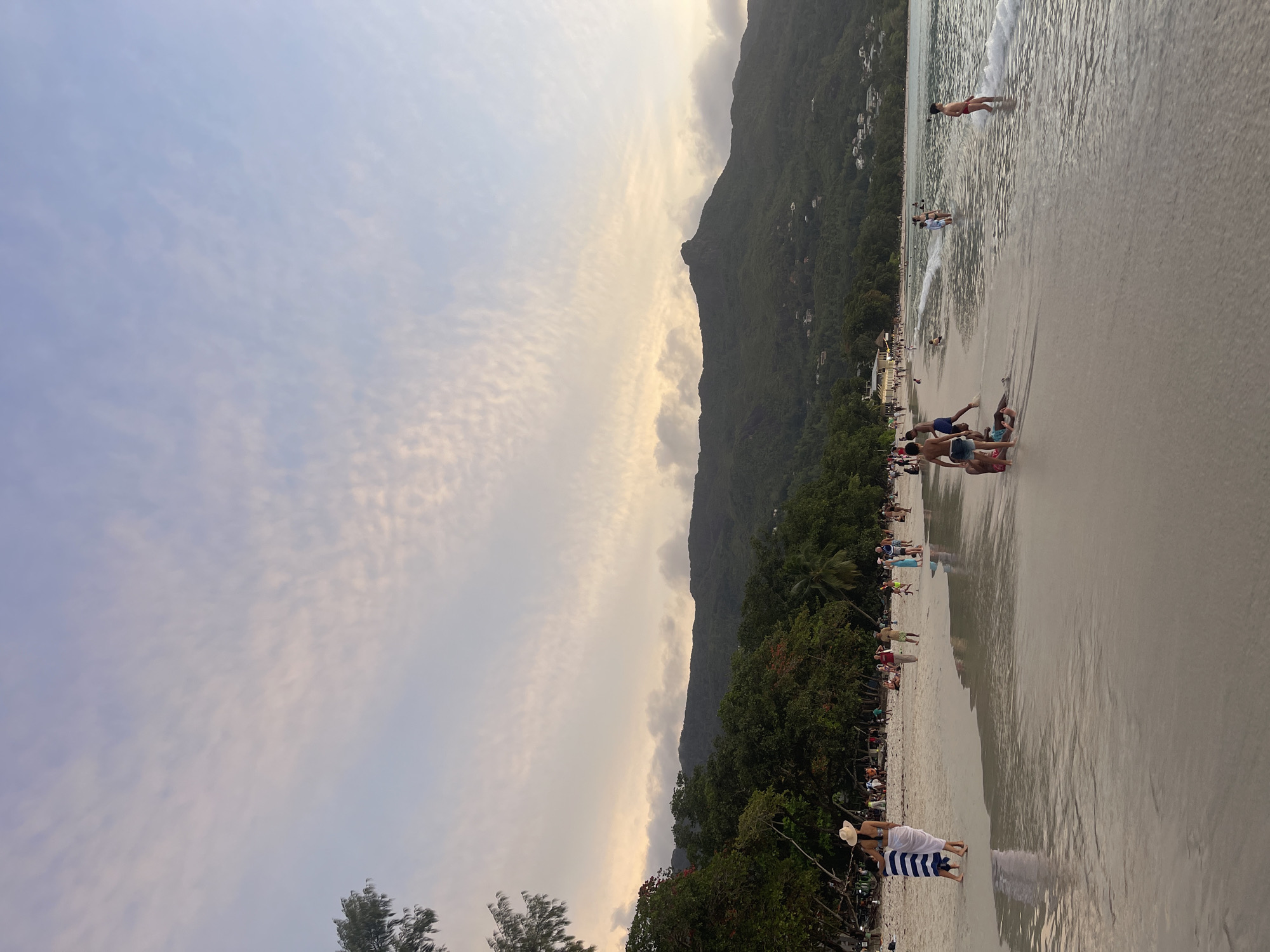This is a vertical, medium-shot, eye-level photograph of Beau Vallon Beach in Beau Vallon, Seychelles, taken at dusk. The sky above is a soft gradient of pale pink and blue, with wispy clouds. In the background, a lush green mountain range rises, dotted with scattered houses and trees. The beach itself is wide and sandy, with the shallow, clear ocean gently lapping at the shore.
Numerous people are visible enjoying the beach. In the foreground, on the left, a woman wearing a white sun hat and a blue and white striped sarong walks away from the camera, accompanied by a child. Further down the beach, a group of children and adults are scattered, some wading in the water, others walking along the shore. A young boy in red swim trunks stands in the shallow water on the right side of the frame. In the middle distance, a line of trees and palm trees separates the beach from the foothills of the mountain. Several structures, possibly restaurants or beach bars, are visible nestled amongst the trees. The overall atmosphere is relaxed and serene, suggesting a pleasant evening at a popular tourist destination.