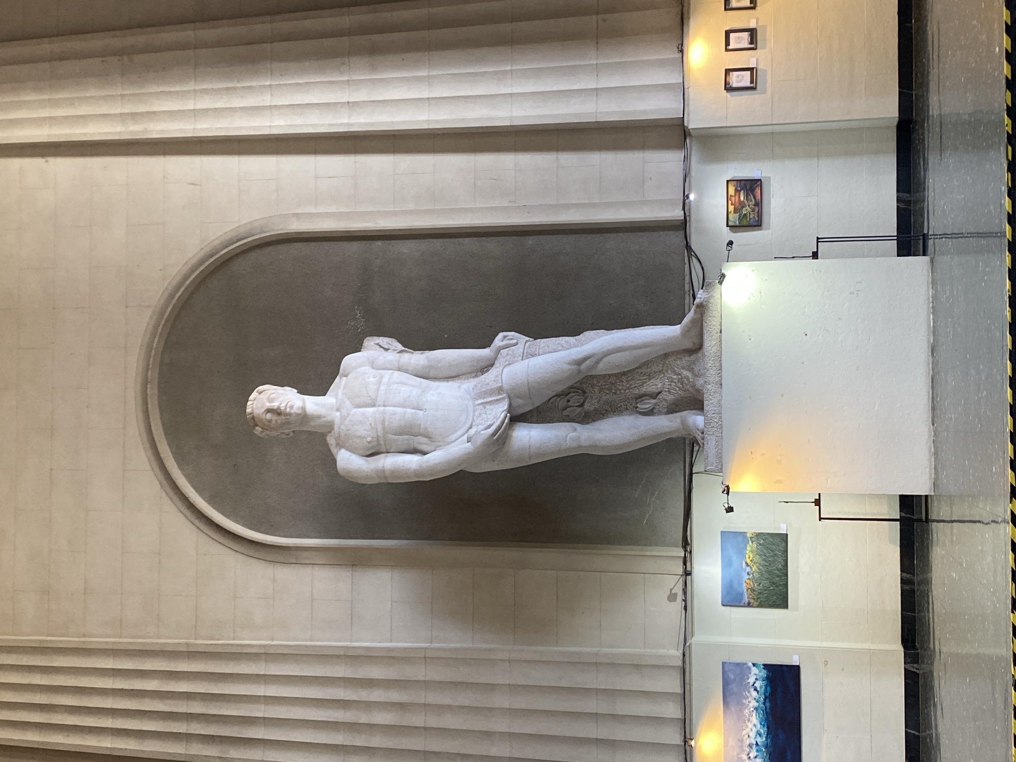 The image shows a large white marble sculpture of a reclining male figure displayed within an arched niche in a hallway or gallery setting. The sculpture is likely of classical or neoclassical style. The walls are composed of light-colored tiles and molding. There are framed artworks hung on the wall to the right of the sculpture, illuminated by spotlights. Some paintings and other art pieces are on display stands. A yellow and black safety barrier is visible on the right side of the image, suggesting the possibility of ongoing renovations or restricted areas. The setting seems to be inside a formal building, likely a museum or cultural center. Based on the sculptures and architectural design, it could be the National Museum of Decorative Arts in Buenos Aires, Argentina.