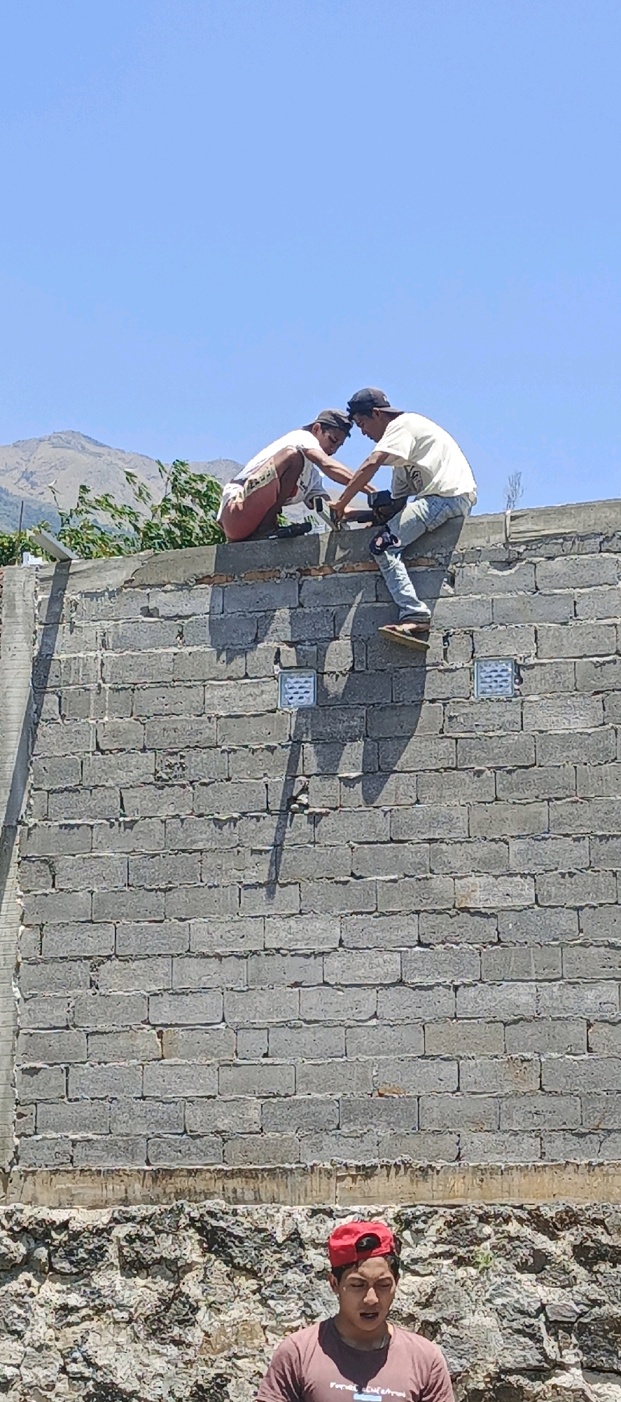 Two men are working on top of a cinder block wall, likely in Jlarem, Indonesia. The man on the left, wearing a white t-shirt and red shorts, is crouched down and appears to be assisting the man on the right. The man on the right, dressed in a white t-shirt, jeans, and a black baseball cap, is standing on the wall and appears to be using a tool, possibly an electric drill, to secure something to the wall. His right foot is resting on a cinder block. Two small, square light fixtures are visible on the wall below them. In the foreground, a young man wearing a red baseball cap and a maroon t-shirt looks upwards, his mouth slightly open. The background shows a clear blue sky, a distant mountain range, and some vegetation. The wall itself is constructed of grey cinder blocks, with some exposed brickwork at the top. The lower portion of the scene is a rocky, uneven embankment. The overall impression is one of ongoing construction work in an outdoor, possibly rural or semi-rural, setting.