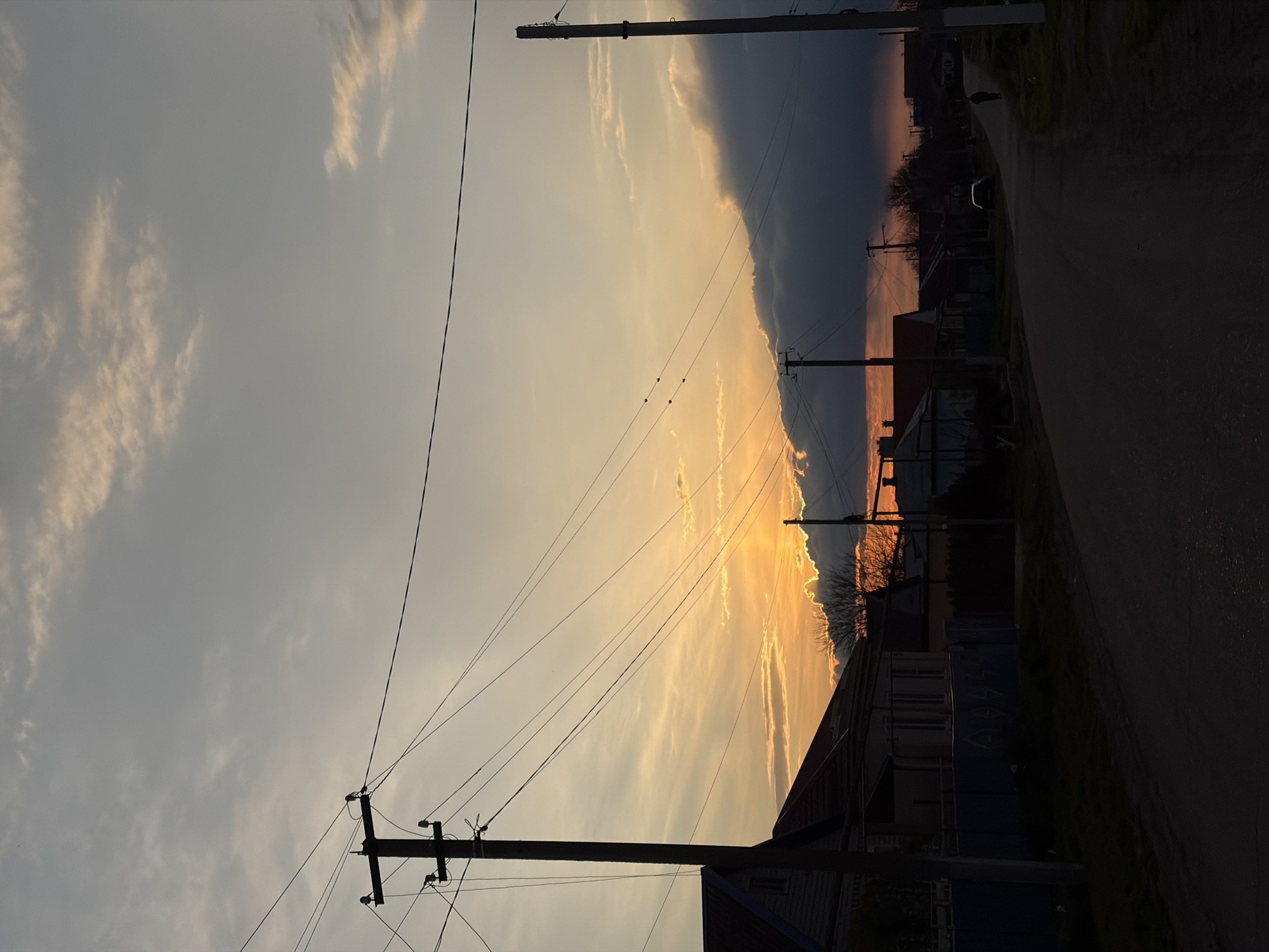 The image depicts a street scene in Novopokrovskaya, Russia at sunset. A paved road runs along the right side of the frame, fading into darkness. The sky is a mix of light blue and orange hues, with dark storm clouds looming near the horizon. Power lines stretch across the sky, supported by wooden poles. Houses and fences line the road.