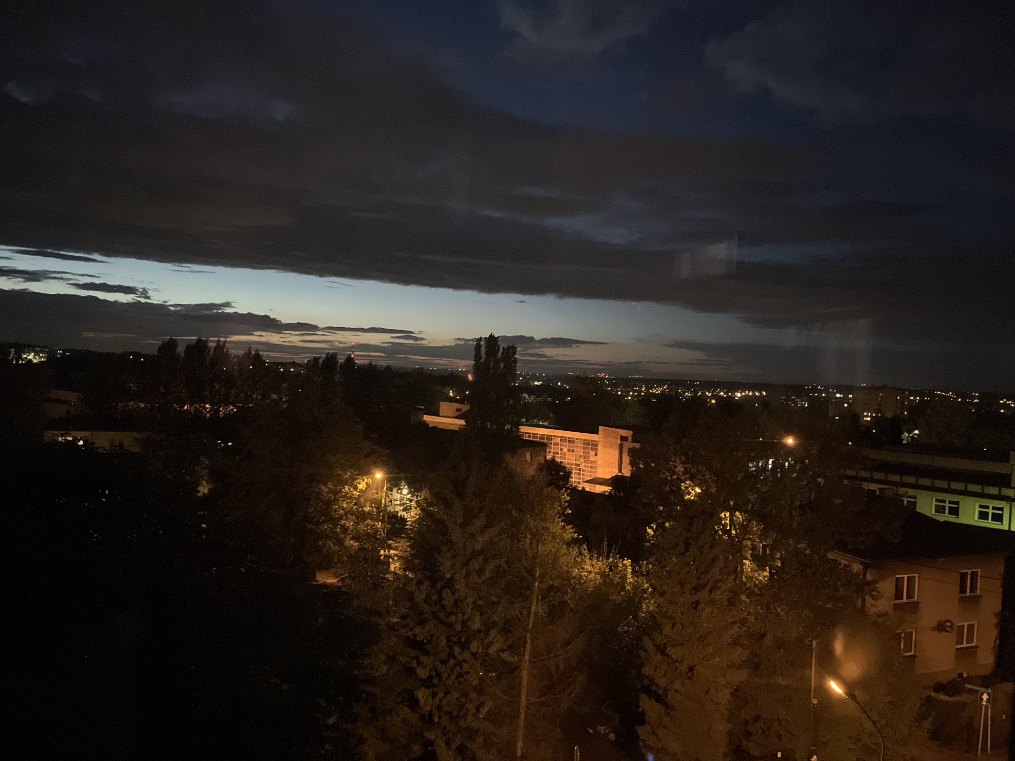 This is a nighttime image of an urban landscape with dark, dramatic clouds in the sky. The foreground is dominated by trees, silhouetted against the dim light. In the midground, there are buildings with illuminated windows, suggesting human habitation. One prominent building appears to be a modern structure with a grid-like facade, possibly an office building or a research facility. Streetlights cast a warm glow on the surrounding trees. The background shows a hazy city skyline dotted with lights, indicating a sprawling urban area. The overall mood is one of a quiet, dusky evening.