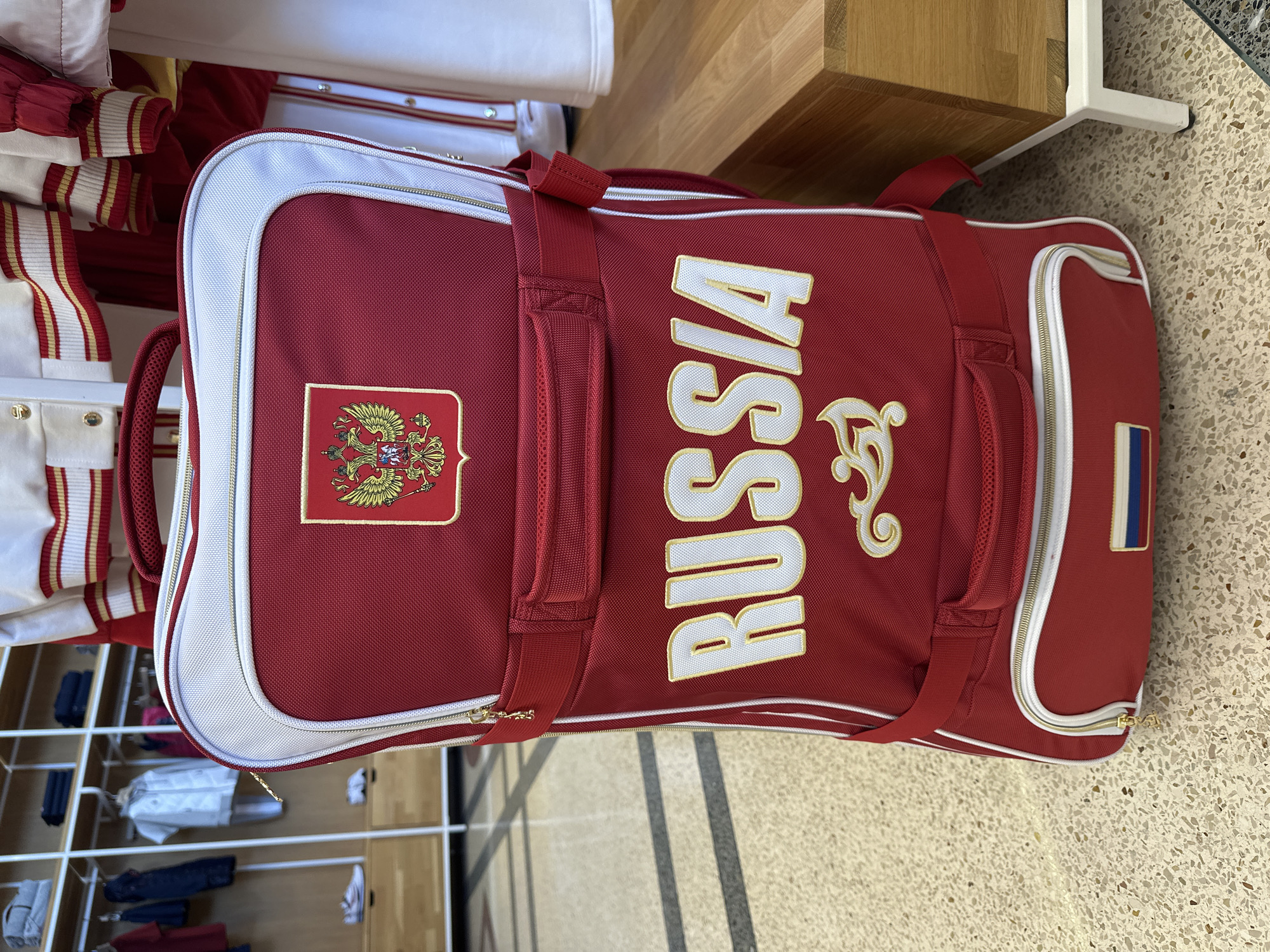 The image displays a large, rectangular, dark red duffel bag, oriented vertically, resting on a speckled, light-colored floor. The bag features prominent white and gold trim along its edges and zippers.
The main surface of the bag is emblazoned with the word "RUSSIA" in large, gold, serif block letters outlined in white. To the left of the text, a patch depicting the golden double-headed eagle of the Russian coat of arms is sewn onto the bag. On the right side, near one end, a small rectangular patch with the colors of the Russian flag (white, blue, red) is visible. The bag has several red fabric handles and straps.
In the background, to the left, there is a rack filled with white and red garments, possibly uniforms or athletic wear, featuring gold striped accents. These clothes appear to be neatly folded or hung. Further back, on the right, some light-colored wooden furniture or shelving is partially visible, along with glimpses of other items on lower shelves. The floor appears to be a terrazzo-like material with varying shades of beige and grey. The overall setting suggests a changing room, locker room, or equipment storage area, possibly for a sports team or official delegation in Sochi, Russia.