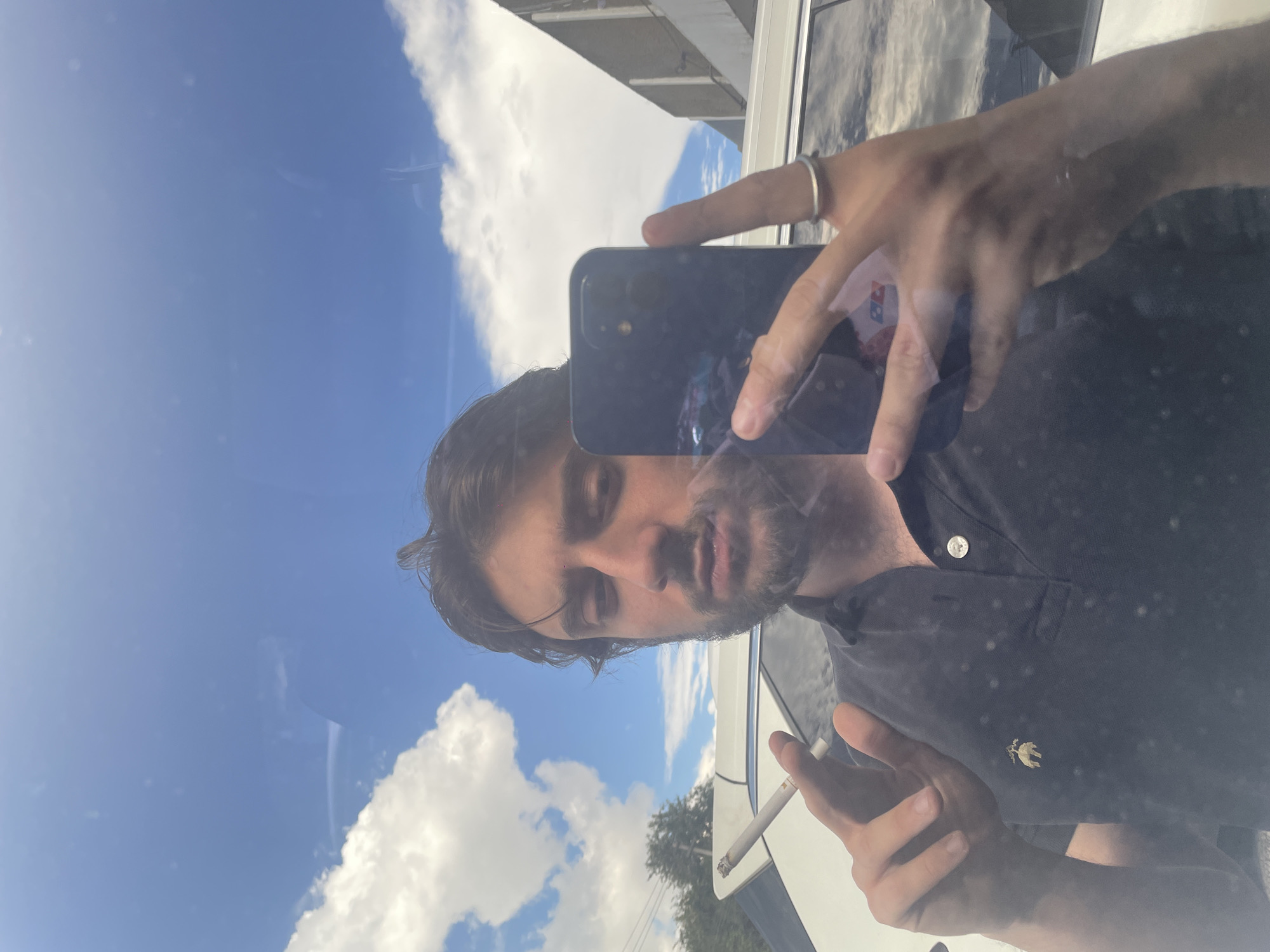 A man stands in front of a white SUV, taking a selfie with his phone. He is wearing a dark blue polo shirt with a golden logo on the left side of his chest. He is holding a lit cigarette in his left hand and has his right hand holding his phone, with a ring on his right index finger. The background shows a bright blue sky with fluffy white clouds, trees, and a building. The scene appears to be outdoors, possibly in the city of Cuautitlán Izcalli, Mexico, given the presence of an SUV and the general appearance of the surroundings.