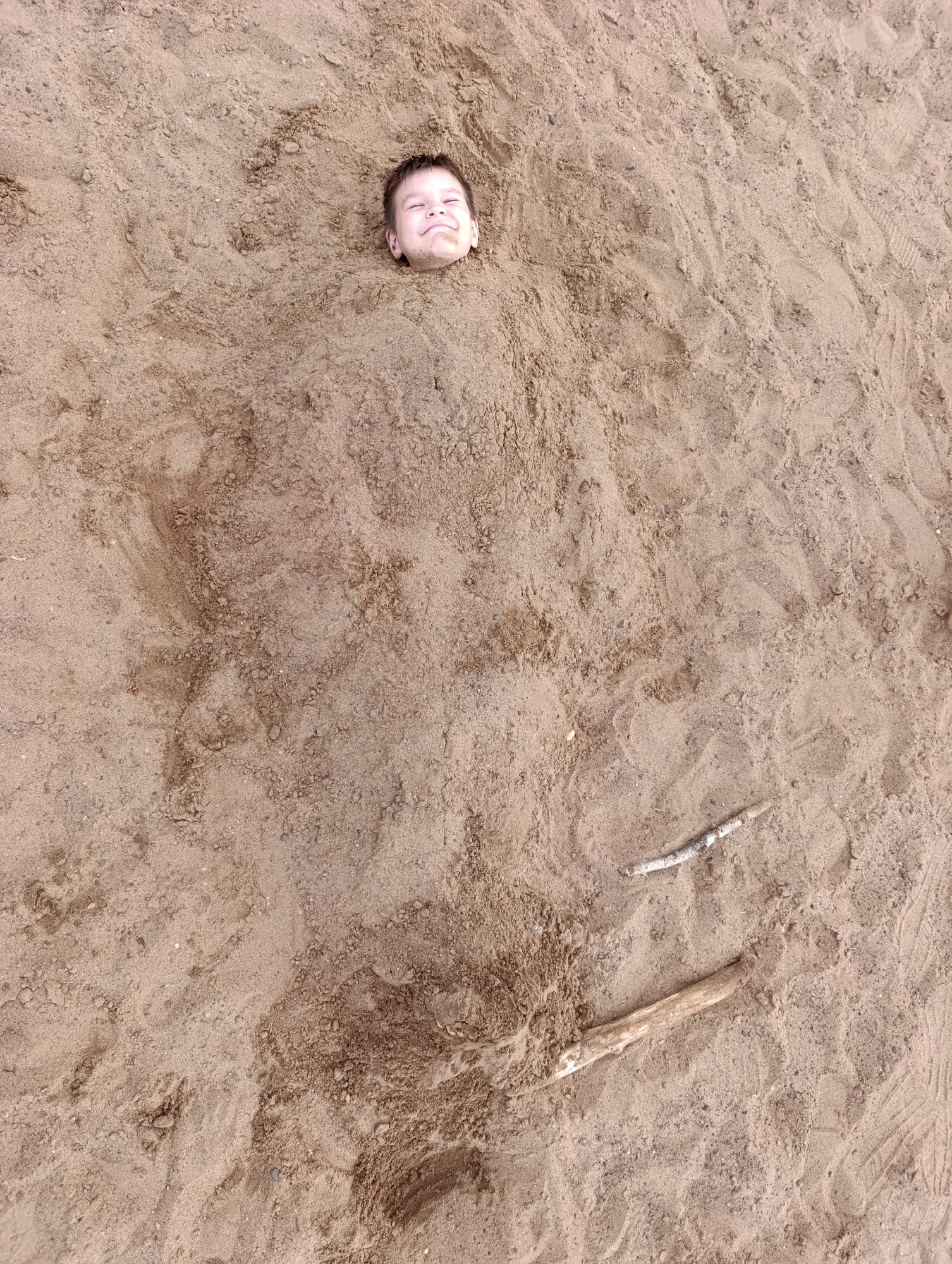 The image shows a young boy buried up to his head in sand. He is smiling with his eyes closed, suggesting he is enjoying himself. The setting appears to be a beach or sandy area, with a few small sticks scattered on the sand. The sand is a light brown color and shows some texture from being disturbed, possibly by footprints. The lighting suggests it is daytime, with the sun casting soft shadows. There is no visible text in the image. The overall mood of the image is playful and lighthearted.