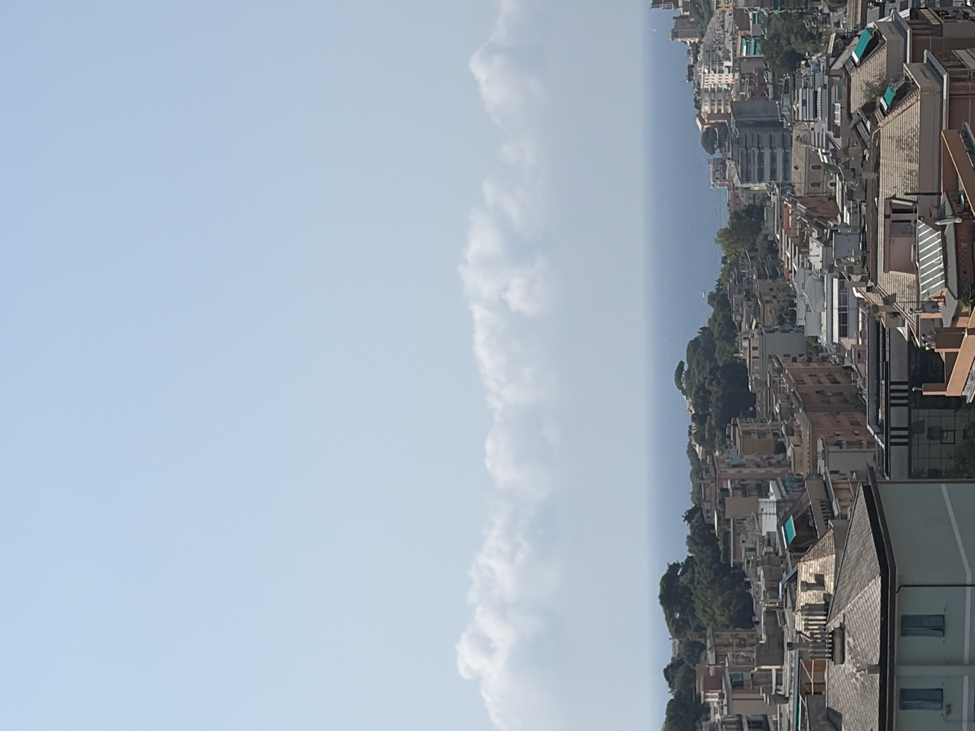 The image presents an elevated view of a densely populated urban area, likely a coastal city in Genoa, Italy. The foreground is dominated by the rooftops of numerous buildings, varying in height and architectural style. Many buildings have terracotta or grey tiled roofs, some with visible chimneys and ventilation systems. Several buildings feature balconies and awnings. A prominent light blue building with dark grey trim and two rectangular windows is visible on the left side of the foreground.
Beyond the immediate rooftops, a dense collection of residential and commercial buildings stretches into the mid-ground. These buildings display a range of colors, including pink, beige, and white, with many having visible windows and architectural details. Green trees are interspersed among the buildings, particularly on the slopes of hills in the mid-ground.
The background consists of the blue expanse of the sea, with a clear horizon line. A band of white, low-lying clouds runs horizontally across the sky above the sea, separating it from the clear, pale blue sky. A faint, distant boat is visible on the water. The overall impression is a bustling Mediterranean city built on hilly terrain, overlooking the sea. There are no people or animals visible in the image.