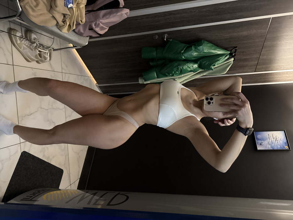 A woman poses in underwear and a sports bra, taking a mirror selfie with her phone. She stands inside what appears to be a tanning salon. Her left arm is slightly bent, holding the phone up to capture the shot. A watch is visible on her wrist. Behind her is a dark wall and a framed certificate. A rack with clothes, shoes, and a green jacket is visible in the background to the left. The floor is tiled. Part of the tanning bed is also visible at the bottom of the frame.