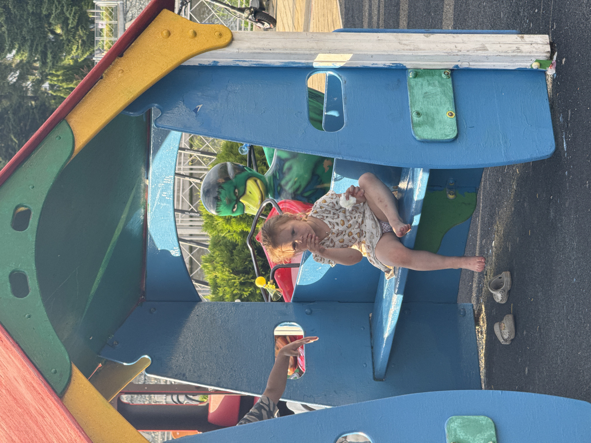 A young girl with blonde hair pulled back sits on a blue wooden bench inside a colorful, multi-level play structure. She wears a white dress with a pattern of small yellow birds and is holding a piece of white food to her mouth with her right hand. Her left arm rests on her leg, and her feet are bare, dangling off the bench. To her left, a person's arm and hand, wearing a striped sleeve, is reaching into the play structure. Behind the girl, a large, green, cartoon-like duck character with angry eyes and a yellow beak is visible. Two pairs of light-colored children's shoes are on the dark, asphalt ground beneath the bench. The play structure, which appears to be made of painted wood, has red and yellow accents on its roof and blue walls. The background shows green bushes and trees, and a white railing suggests an elevated area or walkway. In the far right background, a black electric scooter is parked. The scene appears to be in an outdoor playground.