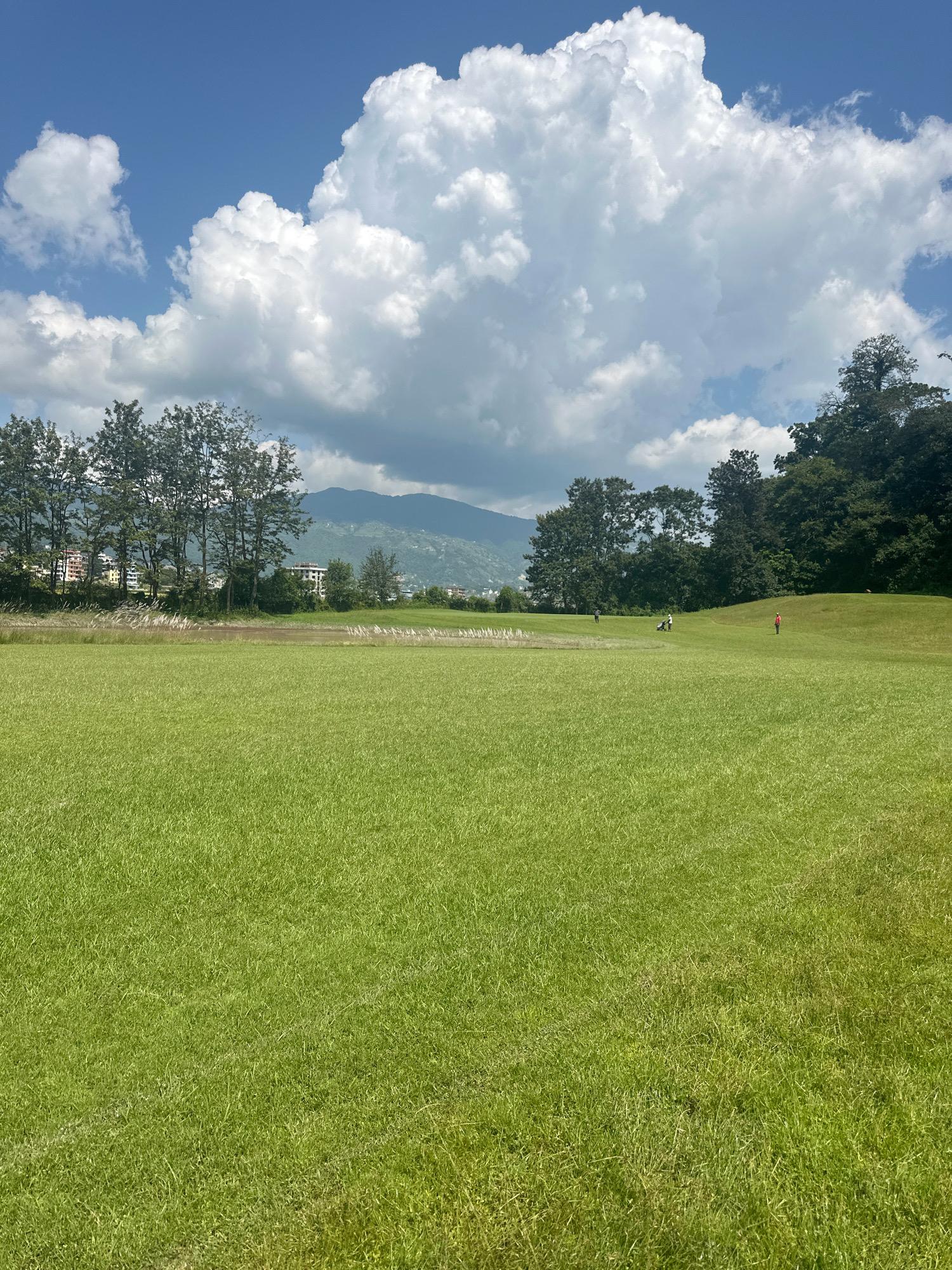The image captures a bright, sunny day at a golf course in Gokarneshwar, Nepal. The foreground is dominated by a vast expanse of meticulously manicured green grass, stretching towards the middle ground. In the distance, several golfers are visible, engaged in their game. One golfer, wearing a red shirt, stands on a gentle slope, while another, with a golf bag, appears to be near a cart. Further back, a cluster of trees borders the golf course, and beyond them, a hazy mountain range can be seen under a sky filled with large, fluffy white clouds. The scene suggests a recreational activity in a picturesque, natural setting within the city.