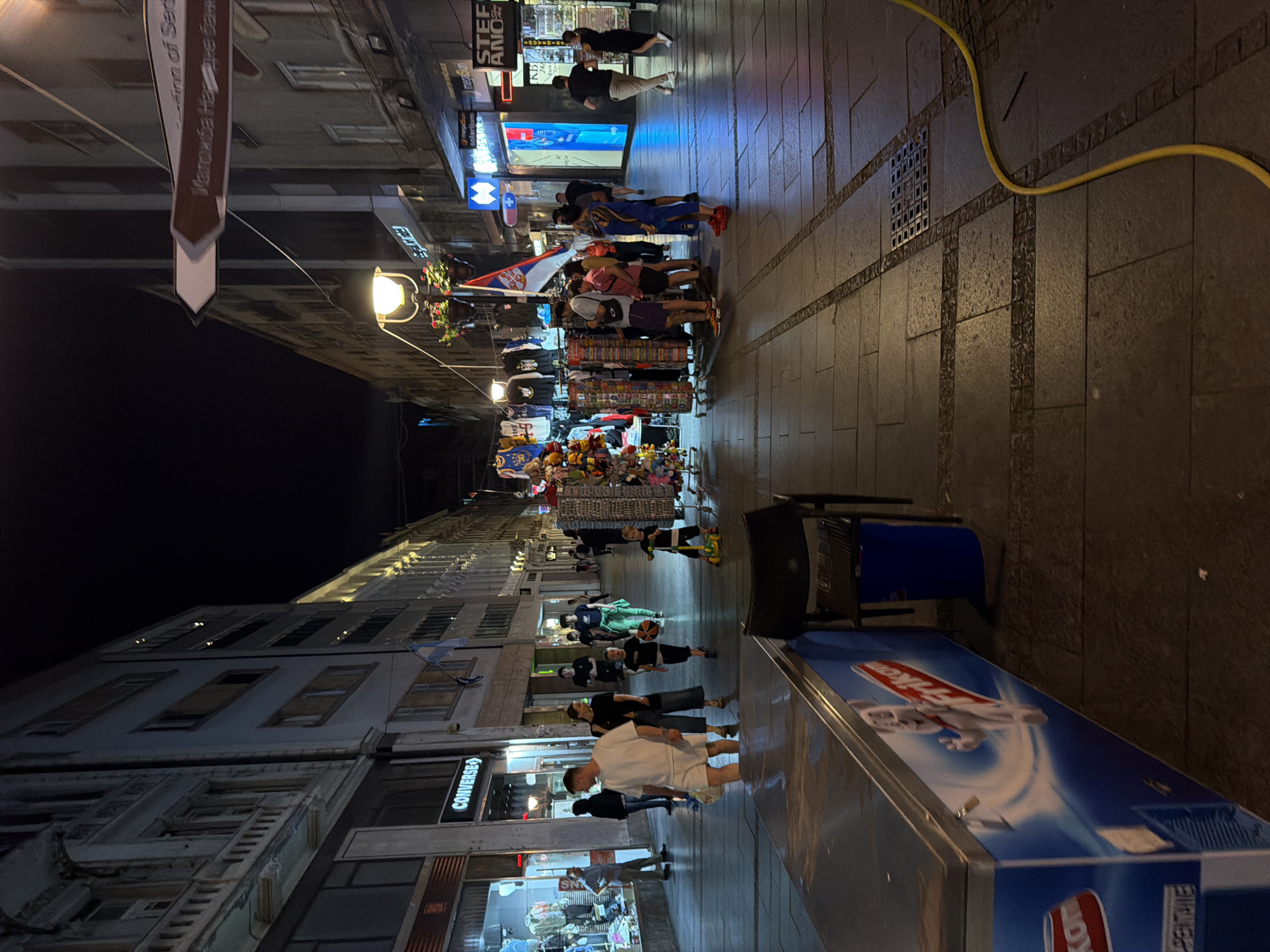 The image depicts a bustling pedestrian street at night in Belgrade, Serbia. The street is lined with multi-story buildings housing various shops and businesses. A prominent sign for "CONVERSE" is visible on one of the buildings. Street vendors have set up stalls displaying merchandise, including clothing and souvenirs. Several people are walking along the street, browsing the shops, and interacting with each other. In the foreground, a refrigerated display case for "Frikom" ice cream is present, along with a blue trash can and a black chair. A yellow hose snakes across the paved walkway, which is illuminated by streetlights and the lights from the shops. The overall atmosphere is lively and commercial, characteristic of a busy urban street at night. A Serbian flag is visible, suggesting the location within Serbia.
