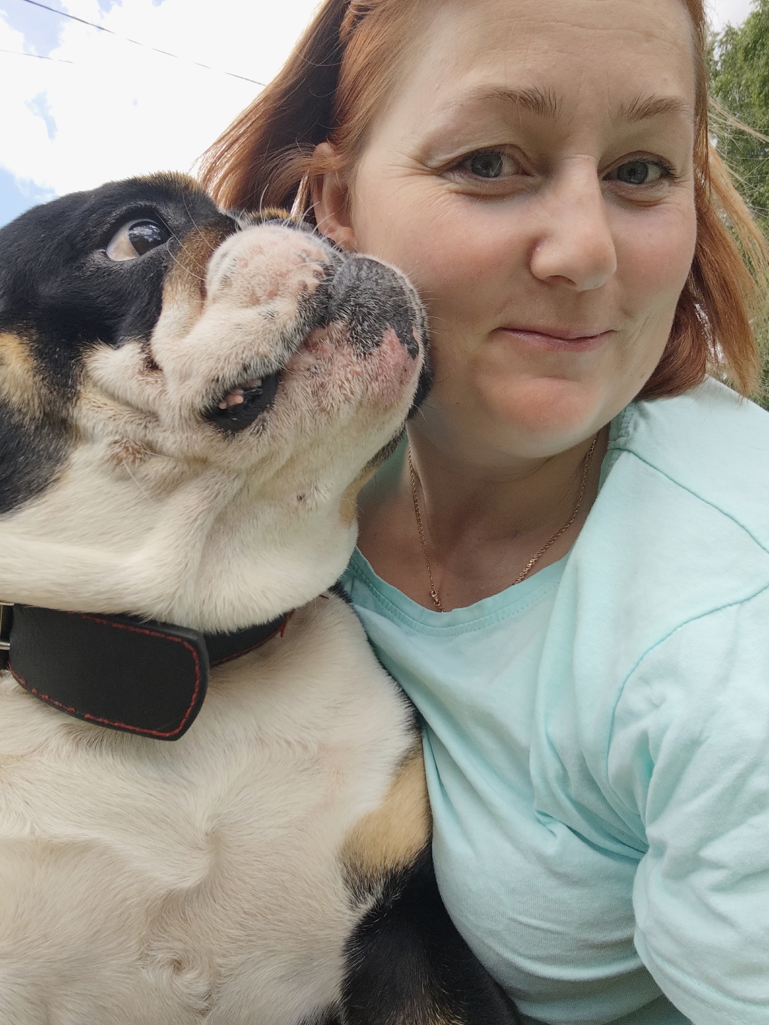 A woman with auburn hair and a light blue t-shirt is taking a selfie outdoors with a bulldog. The bulldog is on the left side of the image, with its face pressed against the woman's cheek. The dog has black, white, and tan markings and is wearing a black collar. The woman is smiling and looking at the camera. The background is blurred, but appears to be an outdoor setting with trees and a sky. The location is the city of Noviy, Russia.