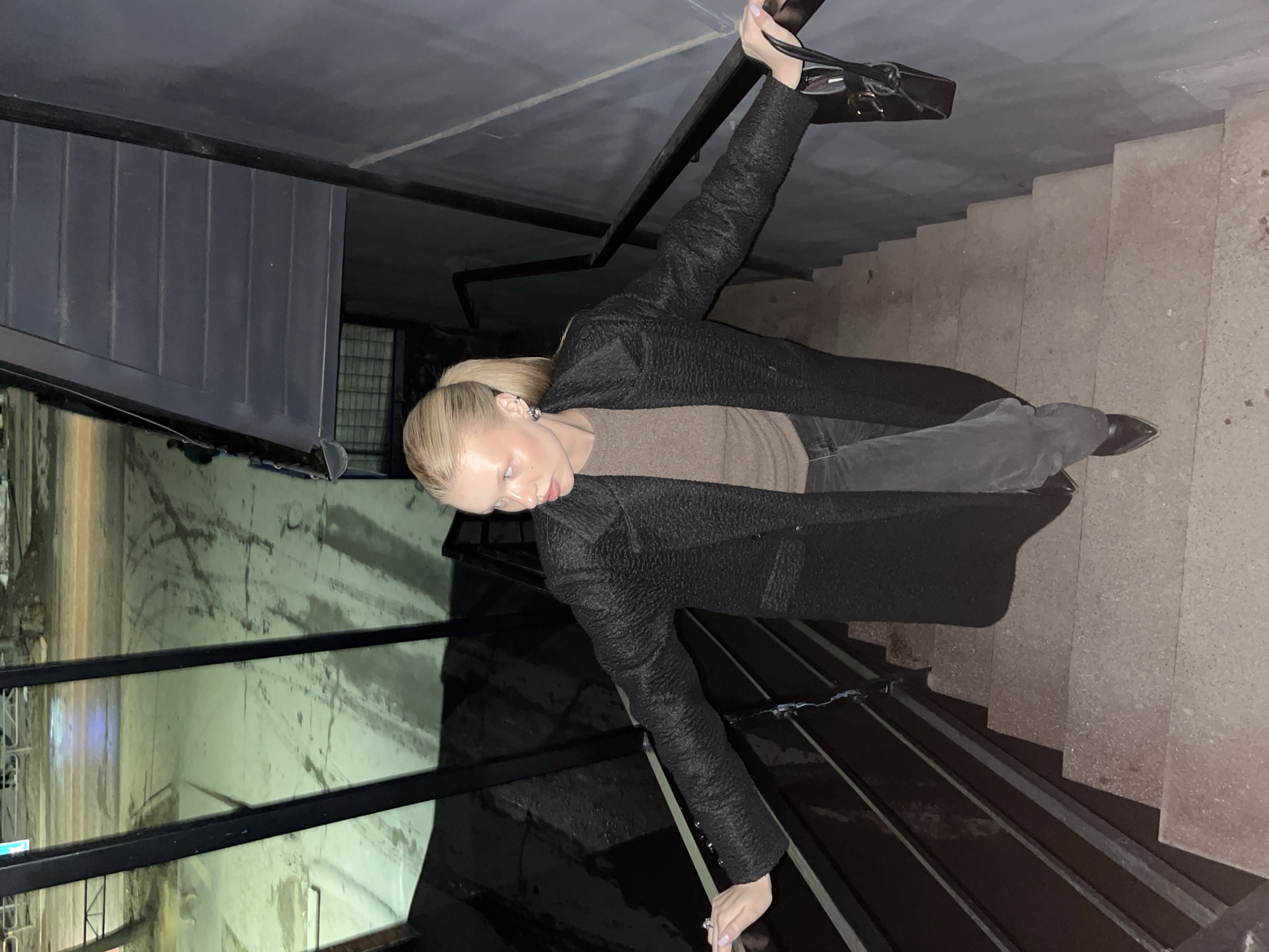 A young, fair-skinned, blonde woman is posing on a metal staircase leading down into what appears to be an industrial or public space. The woman is wearing a brown sweater, dark jeans, and a long, dark-colored coat. She is holding onto the railing with her right hand and holding a black purse with her left hand. The background consists of a concrete wall, a dark metal structure, and the staircase itself. The location is Chaikovsky, Russia.