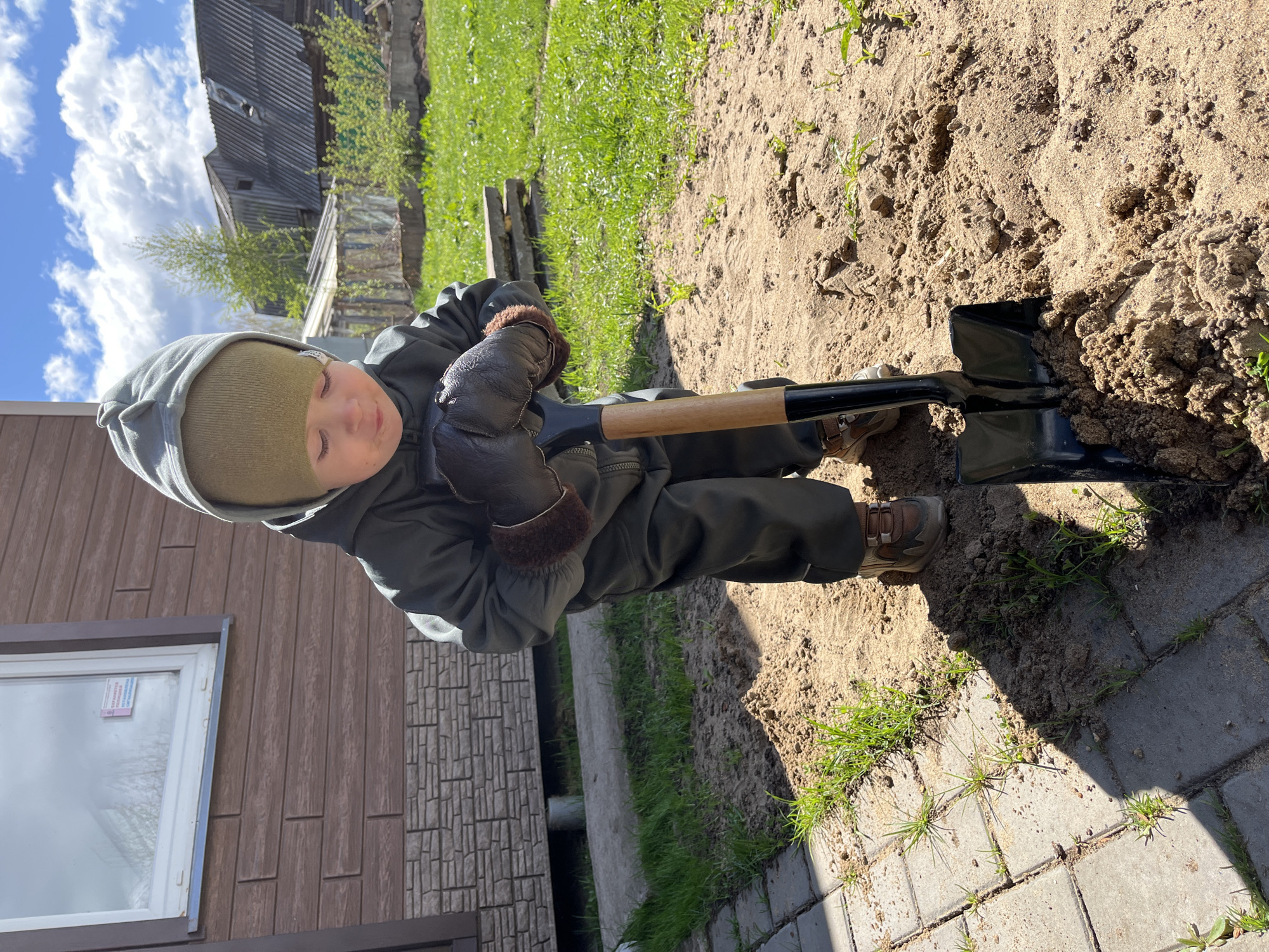 A young child in a green hooded jacket, brown knit cap, and brown leather gloves is digging in sandy soil with a black shovel. The child is also wearing brown sneakers. The shovel is partially embedded in the soil, which is mixed with clumps of dirt and some sparse green grass. The scene includes elements of a yard or garden. Paving stones are visible to the right of the child. The lower corner of a building is seen on the left, featuring brown siding and a window. Another building is in the background. Based on the siding and background, the image is most likely taken in a rural or suburban area, but the exact location in Chaikovsky, Russia is not discernible from the image.