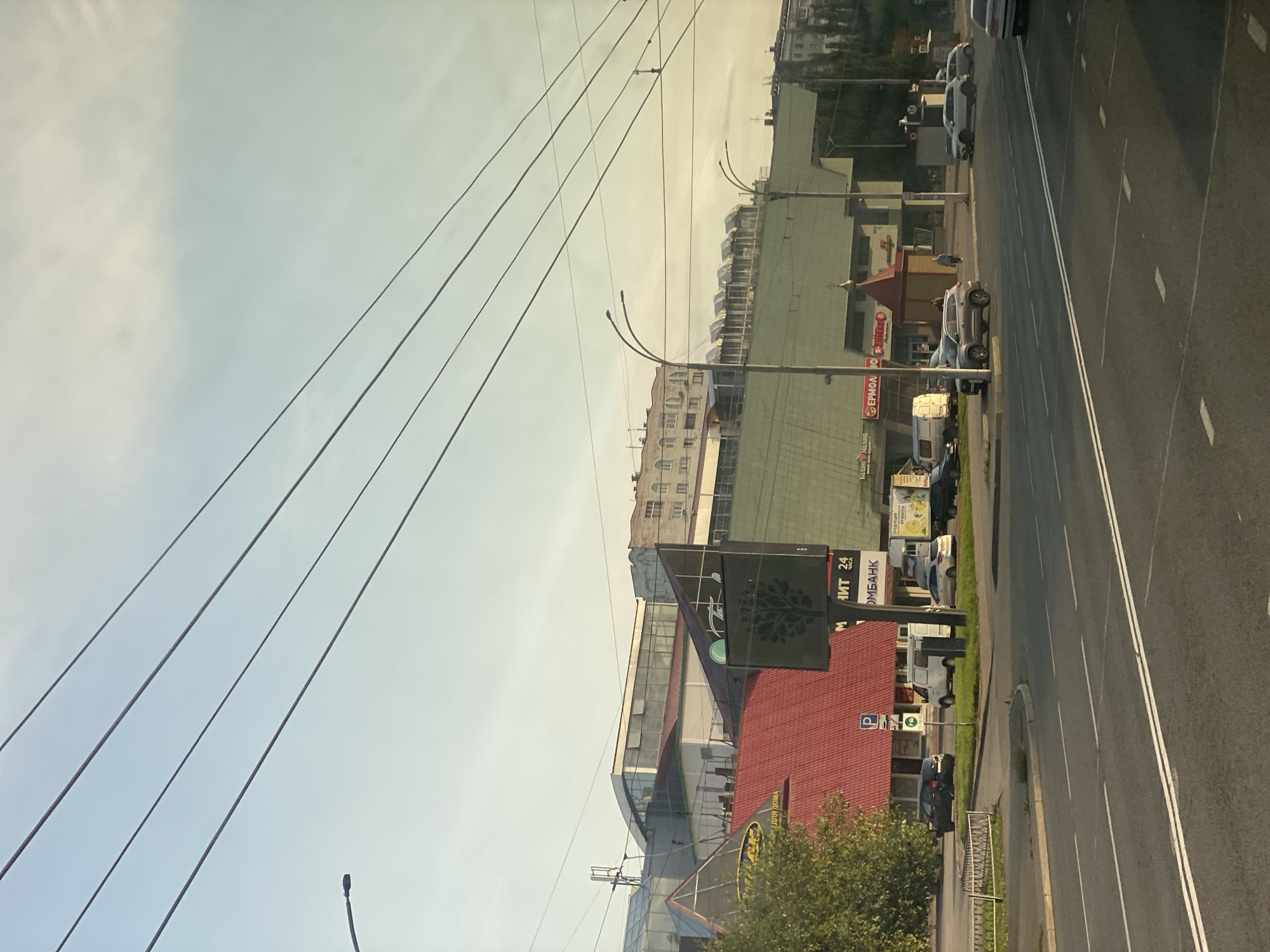 The image depicts a street scene in Kemerovo, Russia, likely in the vicinity of the "Prostor" shopping center. The view is from a moving vehicle, as indicated by the slight blur and the angle of the shot.
Prominently featured is the "Prostor" shopping center, a large, multi-level building with a distinctive green roof and a red, angular facade on the left side. Several vehicles are visible on the road and in parking areas, including a dark sedan, a white van, and a silver SUV. People are present, walking near the shopping center and on the sidewalk. Overhead power lines crisscross the sky, and a street lamp is visible on the left. The sky is a clear, pale blue with a hint of haze, suggesting daytime. The overall environment appears to be an urban commercial district with a mix of modern architecture and infrastructure. Signs for various businesses, including "Промбанк" (Prombank) and "Ермолино" (Ermolino), are visible on the shopping center, further confirming its commercial nature. The road is paved and marked with white lane lines, typical of a city street.