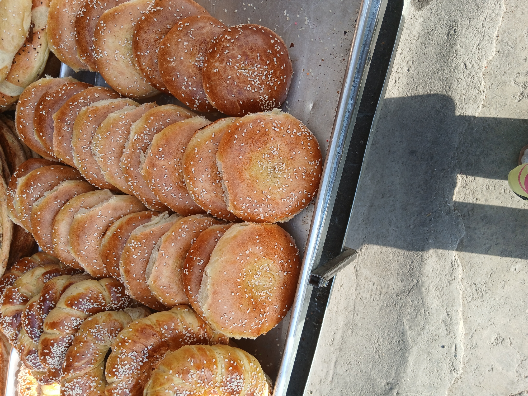 A close-up, overhead shot shows a tray of freshly baked bread, likely at a market in Khankiz, Uzbekistan. The bread appears to be a variety of round, flat loaves and braided rolls, all generously sprinkled with sesame seeds. The bread has a golden-brown crust, indicating it is fresh out of the oven. The tray is made of metal and is placed on a rough, gray surface, possibly a sidewalk or a stall. A shadow cast by a person is visible on the ground, suggesting the image was taken during the day. The overall impression is one of abundance and traditional baking practices.