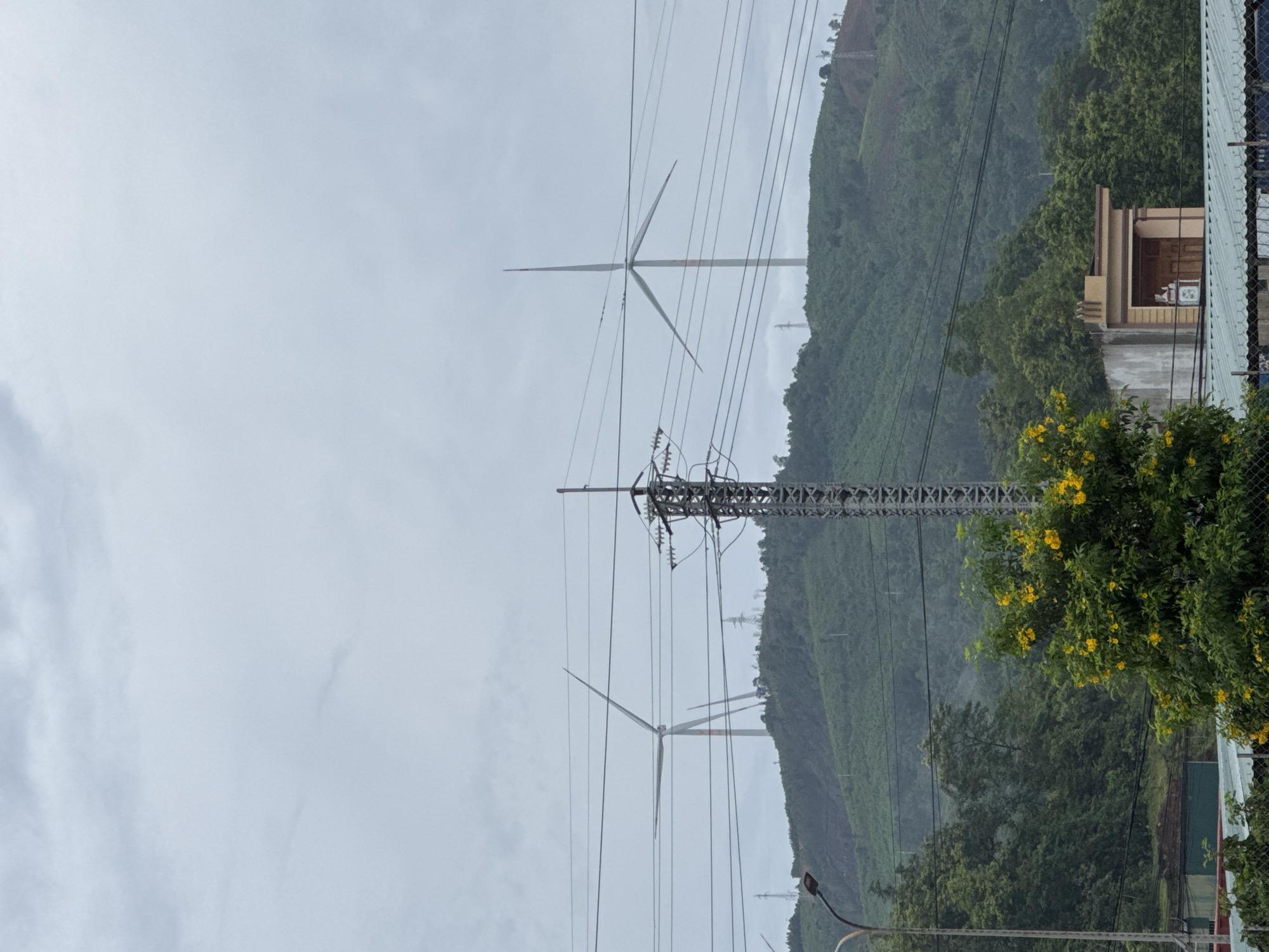 The image depicts a landscape with wind turbines on a hilly terrain under a cloudy sky. In the foreground, there is a transmission tower with multiple power lines. To the left, a street light is visible. A tree with yellow flowers is in the mid-ground, partially obscuring a small building with a covered entrance. The background shows densely vegetated hills with several wind turbines positioned on the higher slopes. The overall scene suggests an area with renewable energy infrastructure and a rural or semi-rural environment. The location is identified as Lao Bảo Commune, Vietnam.