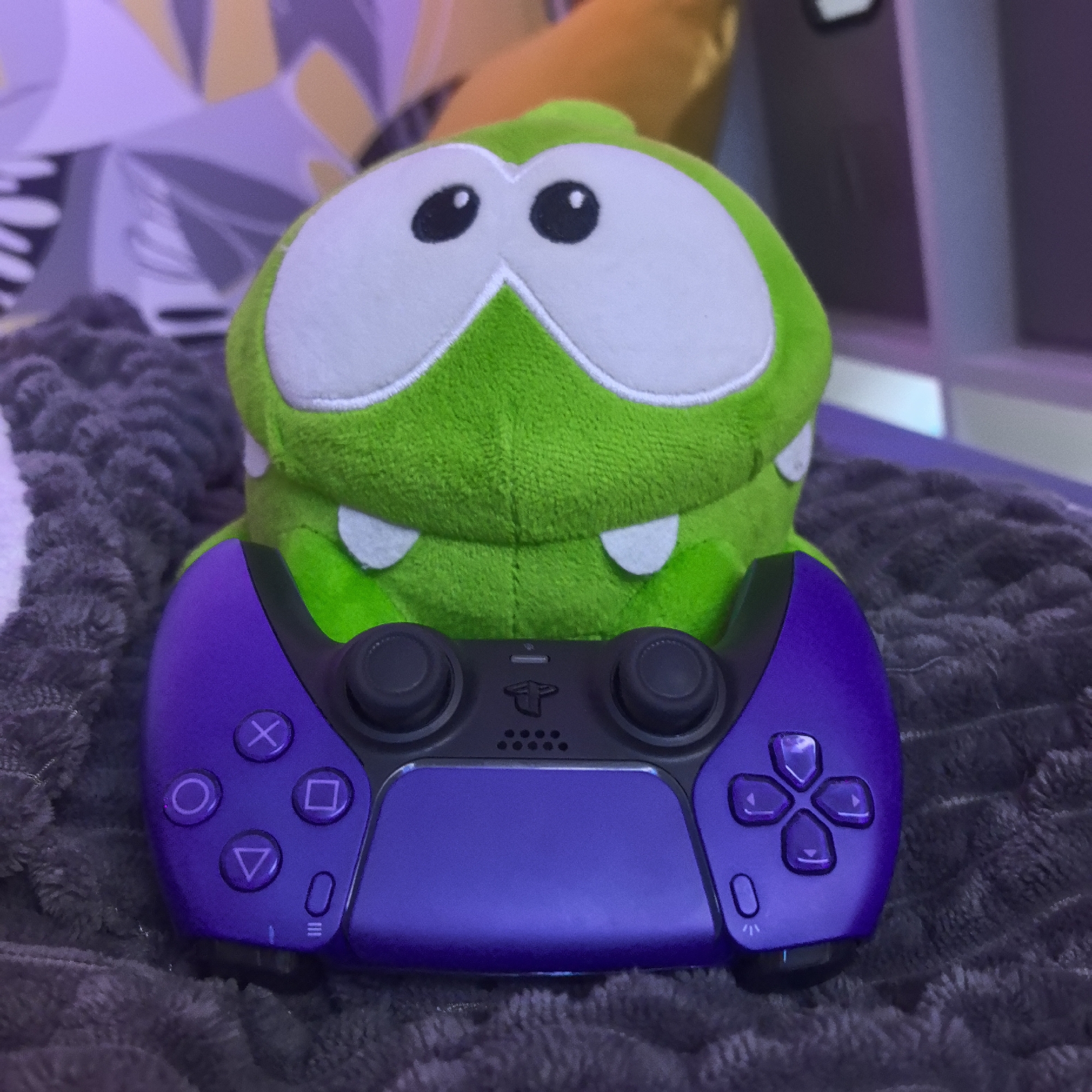 A green plush toy, resembling Om Nom from "Cut the Rope," is depicted sitting on a textured gray blanket, "holding" a purple and black PlayStation DualSense controller. The plush toy features prominent white eyes with black pupils and small white fangs, conveying a playful and endearing expression. The controller, vibrant in its purple hue, shows the PlayStation logo centrally located above the speaker grille and below the share button. The standard PlayStation buttons (Cross, Circle, Square, Triangle) are visible on the left side, while the directional pad (D-pad) is visible on the right. The background is softly blurred, revealing abstract patterns in shades of purple, yellow, and gray, suggesting an indoor setting such as a bedroom or living room. The lighting appears to be artificial, casting a slightly purple ambient glow. The overall scene creates a whimsical and cozy atmosphere, implying the toy is engaging in gaming. No specific time of day or weather conditions are discernible. The image was captured in Khimki, Russia.