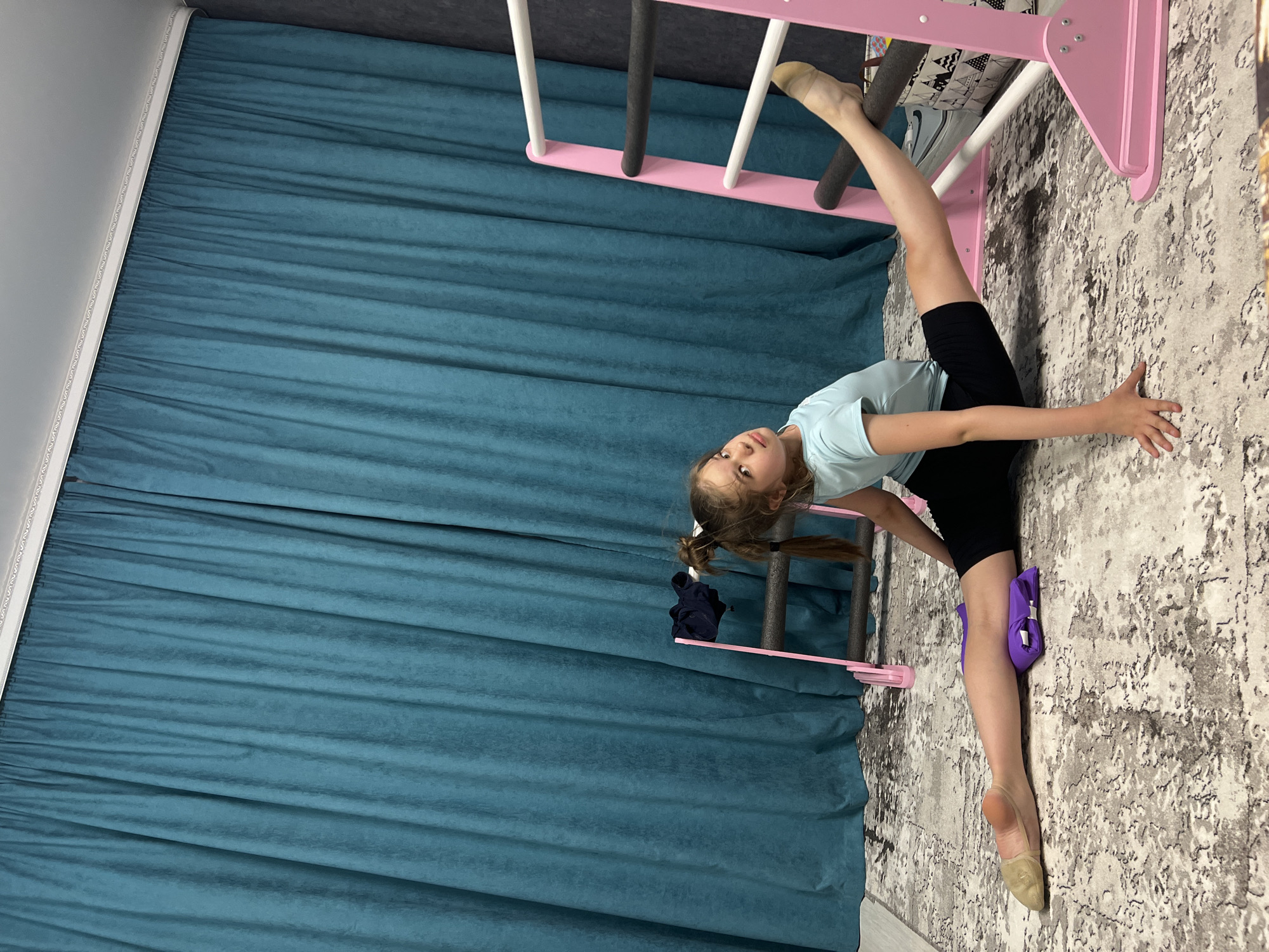A young girl is doing a split in a room with a blue curtain, a beige and white patterned carpet, and a pink gymnastic ladder. She is wearing a light blue t-shirt, black shorts, and ballet shoes. The girl has brown hair styled in pigtails. A purple cushion is positioned under her leg. There is a black t-shirt draped on the ladder. This photograph was taken in Chaikovsky, Russia.