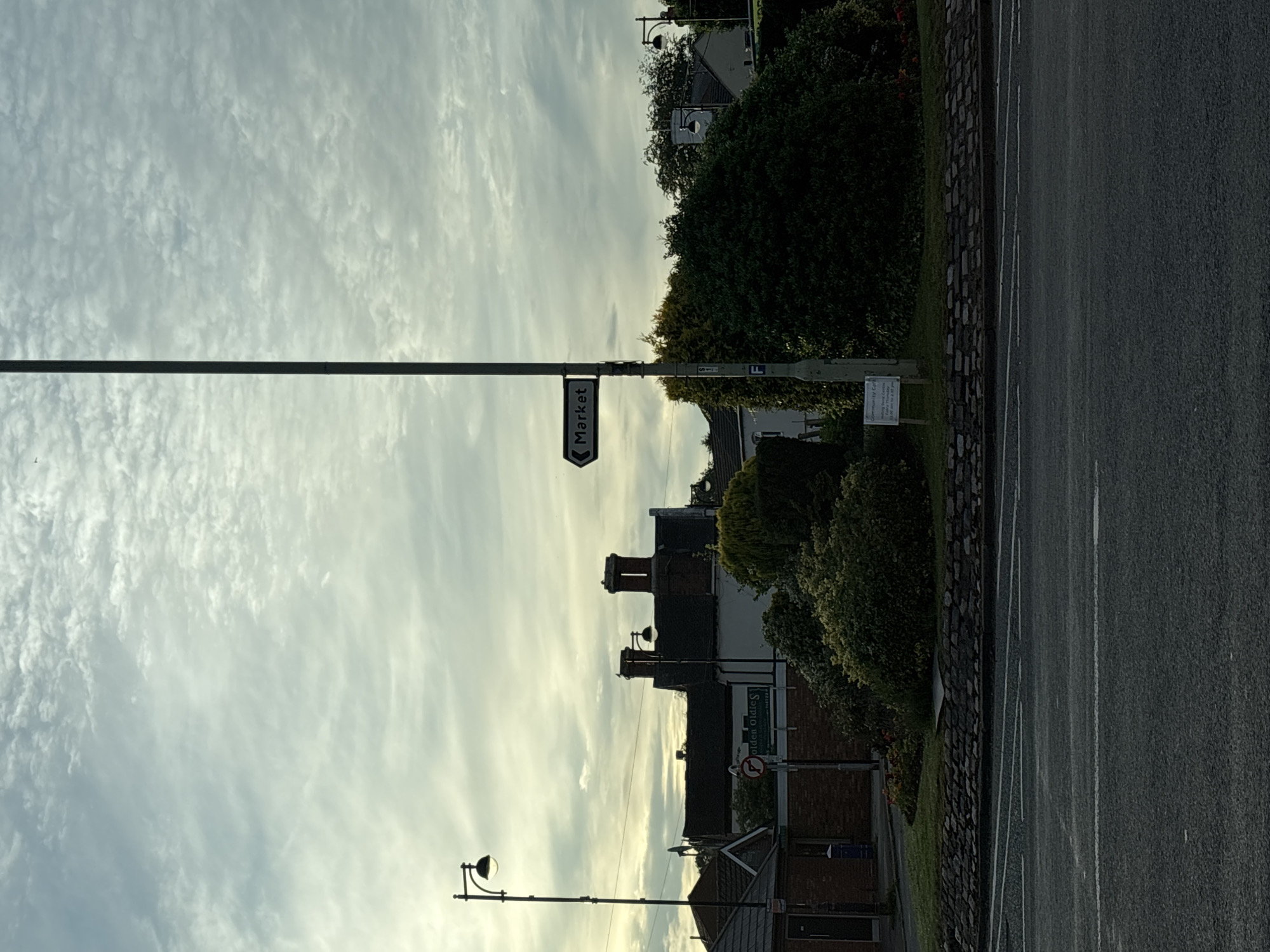 The image shows a street scene in South Staffordshire, United Kingdom, on an overcast day. In the foreground, there is a road with white lane markings. Behind the road, a low stone wall borders a grassy area with some bushes and flowers. A tall lamp post stands on the left side of the image, and another taller lamp post with a directional sign pointing to "Market" is situated more centrally. In the background, there are buildings with red brick chimneys, trees, and more street lighting. The sky is filled with textured clouds. A street sign with a no-entry symbol and another sign with "Community Cafe" and further details are visible.