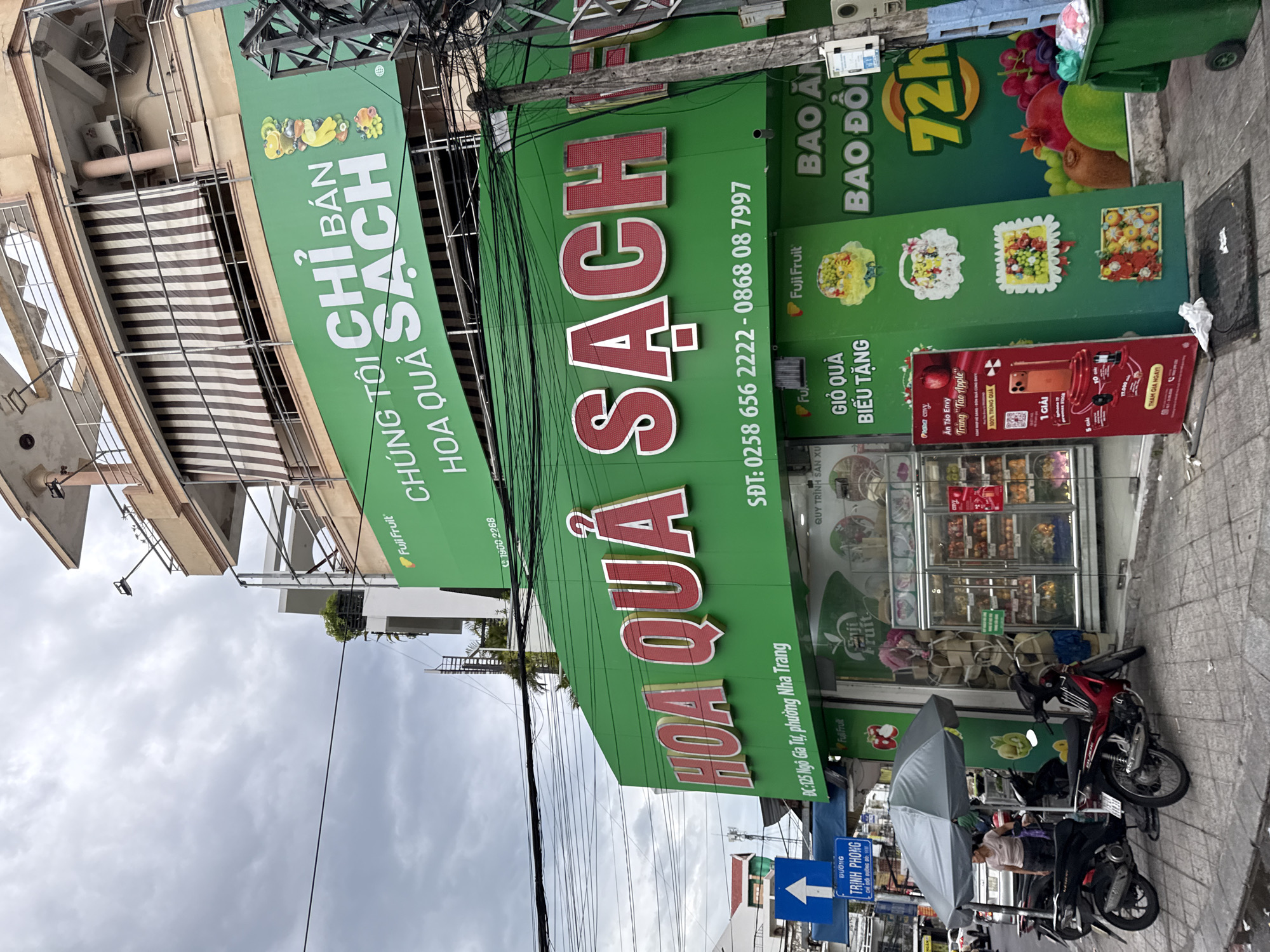 The image shows a street scene in Nha Trang, Vietnam, featuring a Fuji Fruit store. The store has a bright green facade with prominent red and white signage in Vietnamese. The large sign across the top of the store reads "CHÚNG TÔI CHỈ BÁN HOA QUẢ SẠCH," translating to "We only sell clean fruit." Below this, a larger sign states "HOA QUẢ SẠCH," meaning "Clean Fruit." There are also phone numbers and an address displayed: "DC: 125 Ngô Gia Tự, phường Nha Trang." This indicates the store's location at 125 Ngo Gia Tu Street, Nha Trang Ward.
In front of the store, several motorcycles are parked on the sidewalk. A woman in a light-colored t-shirt and dark pants is standing under a large grey umbrella, near a motorcycle. The store interior is visible through glass doors, displaying refrigerated units filled with fruits and other products. A large red advertising stand for a promotion is leaning against the building, showing images of an iPhone and mentioning prizes. To the right of the store, a green trash bin is visible, and large graphics of fruits like grapes, pomegranates, and kiwis are displayed on the store's exterior. The sky is overcast, suggesting a cloudy day. The overall scene depicts a commercial area in Nha Trang with a focus on a fruit store and its offerings.