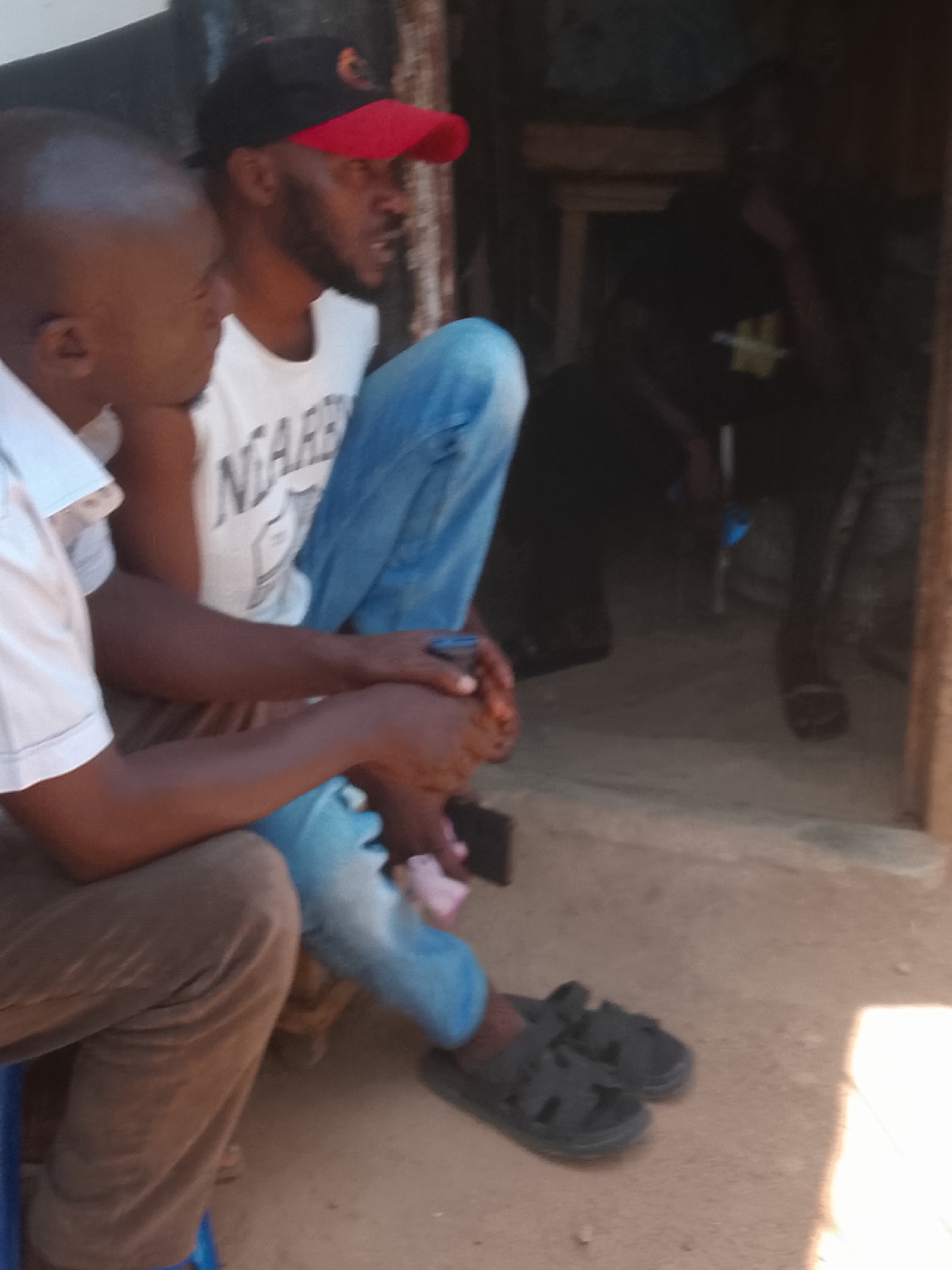 Gwong, Nigeria. Two men sit outside a dimly lit building. One is wearing a red and black baseball cap, a white t-shirt, and blue jeans. The other man is bald, wearing a white short-sleeved shirt and brown pants. Both are wearing sandals. A third person is sitting further inside the building.