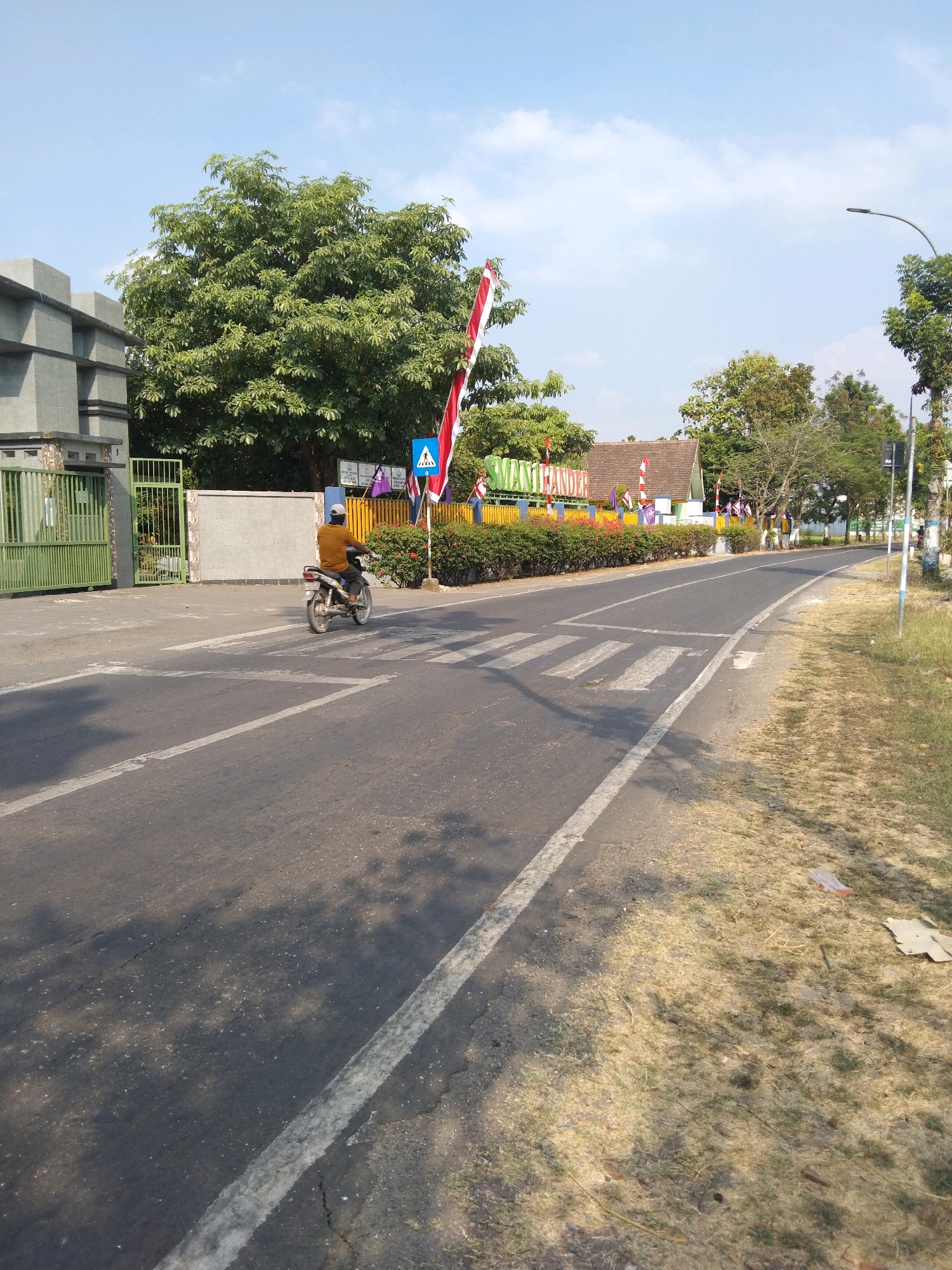 The image captures a street scene in Growok, Indonesia, under a clear blue sky. In the foreground, a motorcycle is crossing a pedestrian crosswalk on an asphalt road. The motorcycle is silver and white, with a rider wearing a brown shirt and dark pants. To the left of the road, there is a green gate and a portion of a building with grey concrete walls. Beyond the gate, a large green tree stands tall. Further down the road, a building with a red-tiled roof is visible, adorned with colorful banners and flags, including the Indonesian flag. The road is bordered by dry grass and vegetation on the right side. A lamppost is visible in the distance. The overall environment appears to be a suburban or semi-urban area, possibly near an educational institution given the signage and flags.