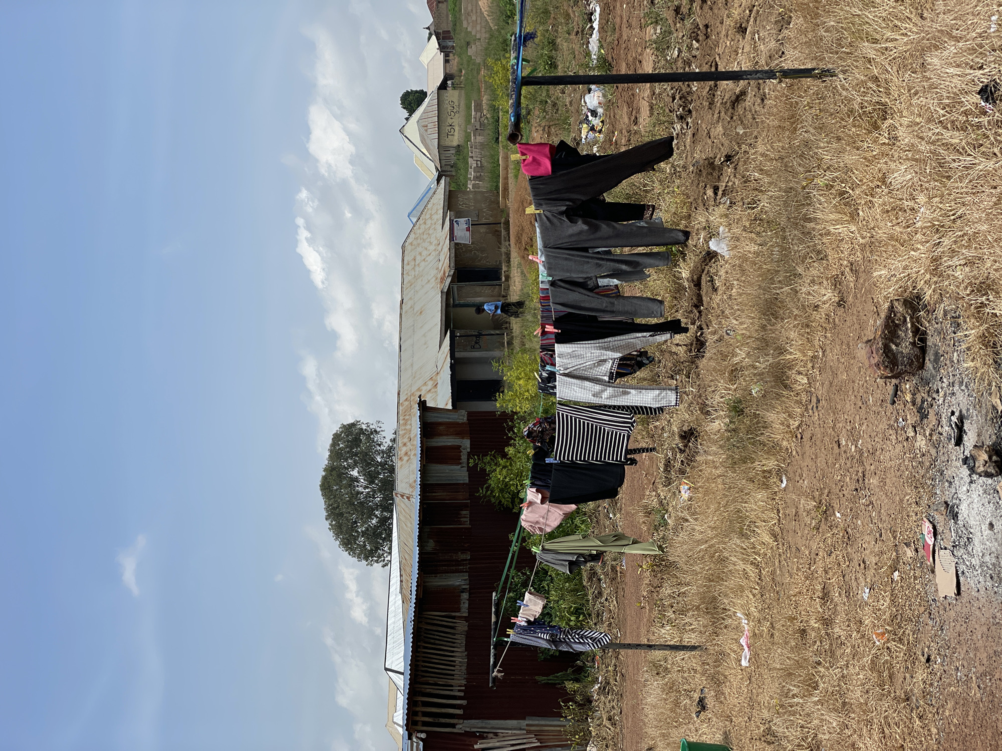 The image depicts an outdoor scene in Durumi, Nigeria, featuring a clothesline laden with various garments drying in the sun. The clothesline is strung between two posts, with clothespins holding the laundry in place. In the background, several buildings with corrugated metal roofs are visible, suggesting a residential or community area. The ground is covered in dry grass and dirt, with scattered debris. The sky is partly cloudy. A person, possibly a young boy or girl, is seen walking near one of the buildings in the background. The overall environment appears to be rural or semi-urban, with a focus on everyday life activities.