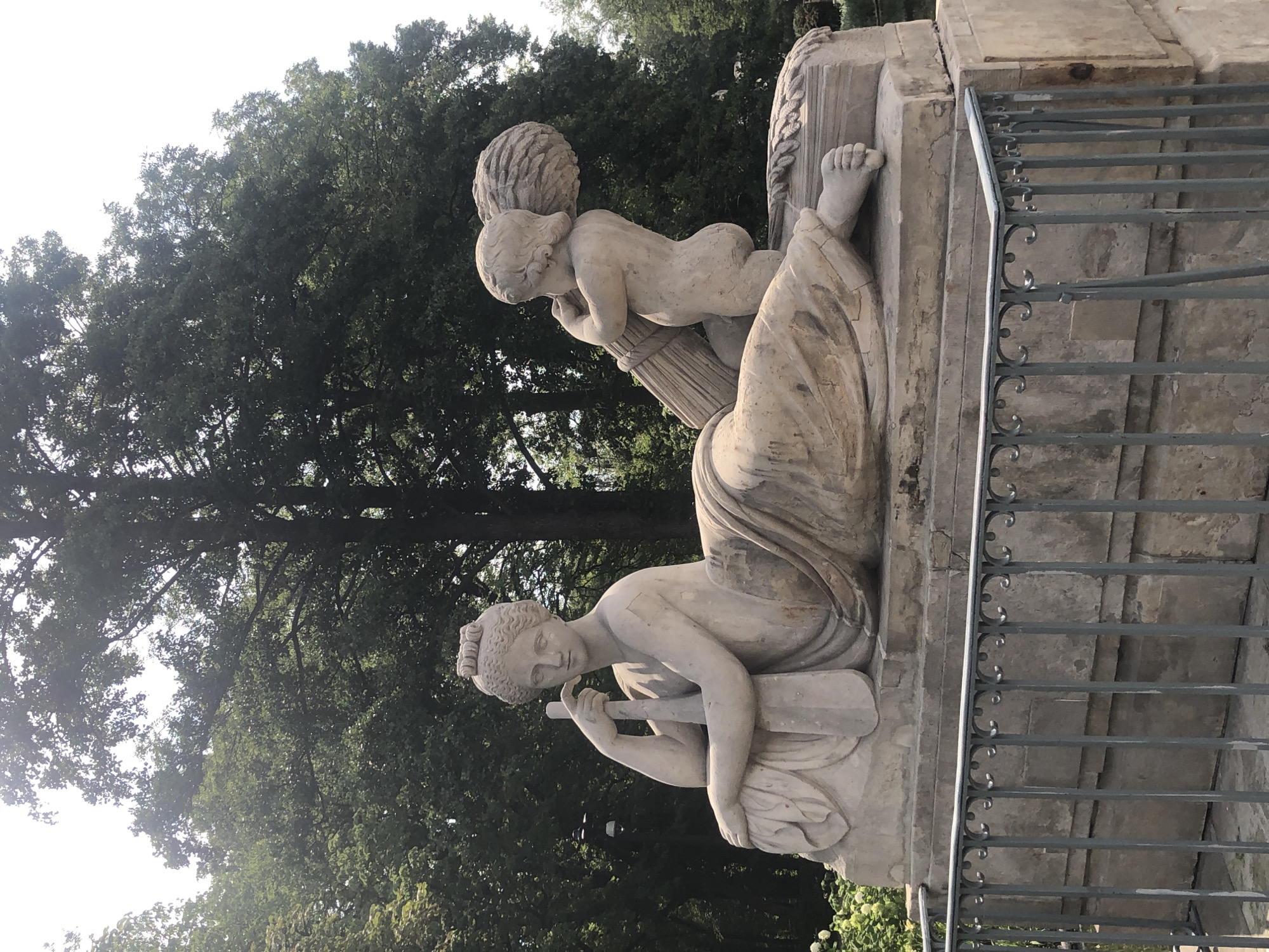 The image shows a stone sculpture of a reclining woman and a cherub in a park. The woman is seated, leaning on her left arm, and holding a staff. She is wearing a draped garment. The cherub is kneeling and holding a bundle of wheat. The sculpture is on a stone pedestal. In the background, there are trees. The sculpture is located in Łazienki Park in Warsaw, Poland.
