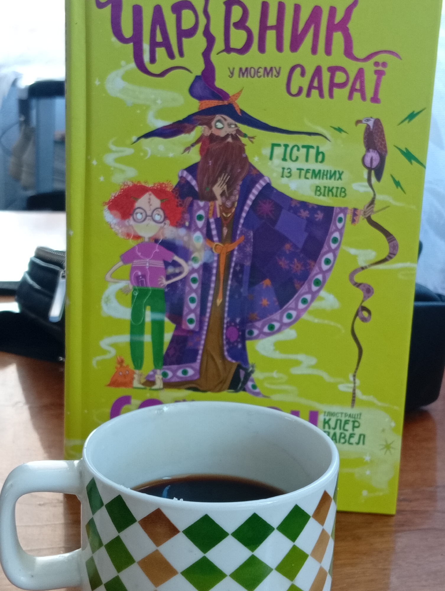 A high-angle medium shot captures a coffee mug in the foreground with a book positioned behind it. The mug, decorated with a green and brown checkered pattern, is filled with dark liquid, presumably coffee. The book, with a vibrant lime-green cover, features an illustration of a wizard and a girl. The wizard, depicted with a long beard and wearing a purple robe adorned with eye motifs, is accompanied by a bird perched on a stick. The girl, with curly red hair and glasses, stands next to the wizard. The book's title, "ЧАР ВНИК У МОЄМУ САРАЇ," is prominently displayed in purple lettering. Text on the book also reads "ГІСТЬ ІЗ ТЕМНИХ ВІКІВ" and "ІЛЮСТРАЦІЇ КЛЕР АВЕЛ". The scene appears to be set on a wooden surface, possibly a desk or table. The background shows a blurred office-like environment with a glimpse of furniture. The lighting is moderate, and the overall image quality is good.