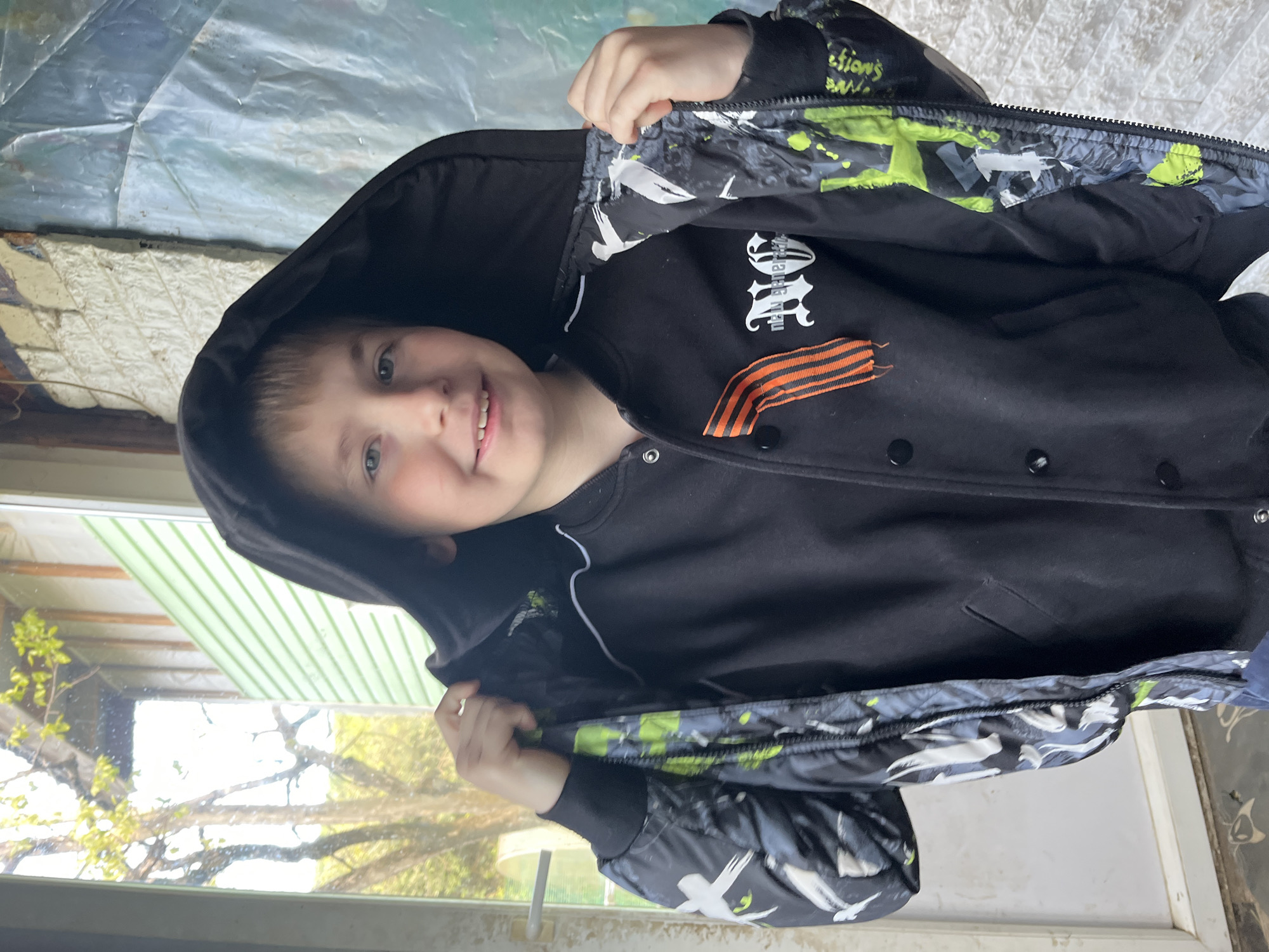 A young boy with light brown hair and blue eyes is captured indoors, smiling and posing for the camera. He appears to be in early childhood and expresses happiness and playfulness. The boy is wearing a black hooded jacket, which he holds open with both hands, revealing its inner lining and the garment underneath. The jacket's lining and sleeve cuffs feature a distinctive grey and lime green camouflage-like pattern, interspersed with white silhouettes resembling birds.
Beneath the jacket, the boy wears a black garment, likely a t-shirt or sweatshirt, displaying stylized white text that reads "NEw Generation," with another word partially visible above it. A prominent orange and black striped ribbon, characteristic of the St. George ribbon, is pinned or sewn onto the black garment on his chest.
The setting appears to be a room illuminated by natural light, likely from a window. Outside the window, green foliage and tree branches are visible, suggesting an outdoor natural environment. Above the boy's head, an indoor wall or surface is present, along with a translucent, crinkled material, possibly plastic sheeting. The scene is set during the daytime. The presence of the St. George ribbon, combined with the location context of Vesyoloye, Russia, suggests a cultural or patriotic theme.