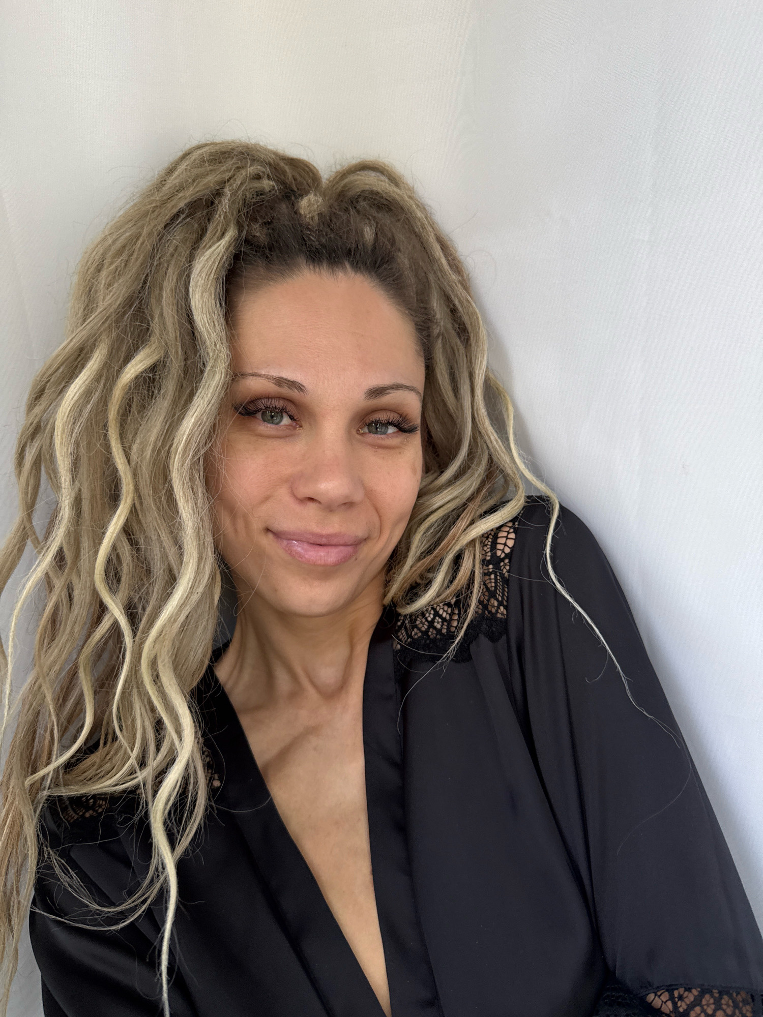 A close-up portrait shows a woman with blonde, wavy hair, wearing a black robe with lace trim. She has green eyes and light skin, and she is smiling slightly. The background is a plain white wall, creating a simple and intimate setting. The image is lit naturally, highlighting the woman's features and the texture of her hair and clothing. The overall impression is one of relaxed femininity. This scene is not located in Rusanovo, Russia.