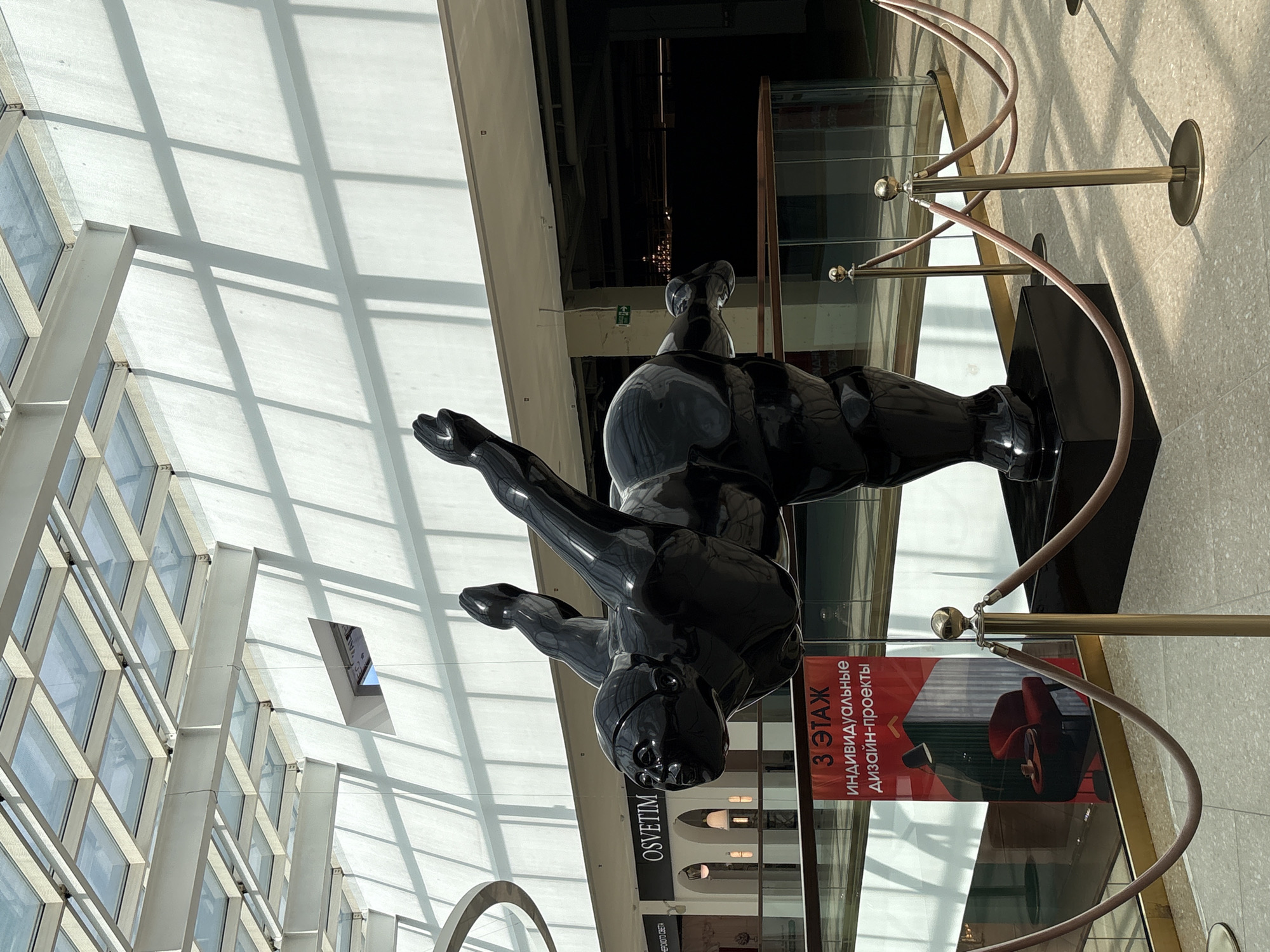 The image shows a large, black, glossy sculpture of a human figure in a dynamic pose, positioned in the atrium of a modern shopping mall in Saint Petersburg, Russia. The sculpture appears to be made of polished metal or plastic and depicts a person with exaggerated musculature, with limbs extended in a seemingly acrobatic or dance-like movement. The figure is viewed from the side, with its head turned towards the viewer.
The environment is brightly lit by natural light filtering through a glass ceiling, creating strong geometric shadows on the white walls of the mall. The background reveals a multi-level interior with shops visible on the upper floors. A sign in Russian, advertising "3RD FLOOR INDIVIDUAL DESIGN PROJECTS" with an image of a modern armchair and lamp, is prominently displayed near the sculpture. There are glass railings with gold stanchions and rope barriers cordoning off the area around the sculpture. The floor is paved with light-colored tiles. No people are visible in the immediate vicinity of the sculpture, but the setting suggests a public indoor space.