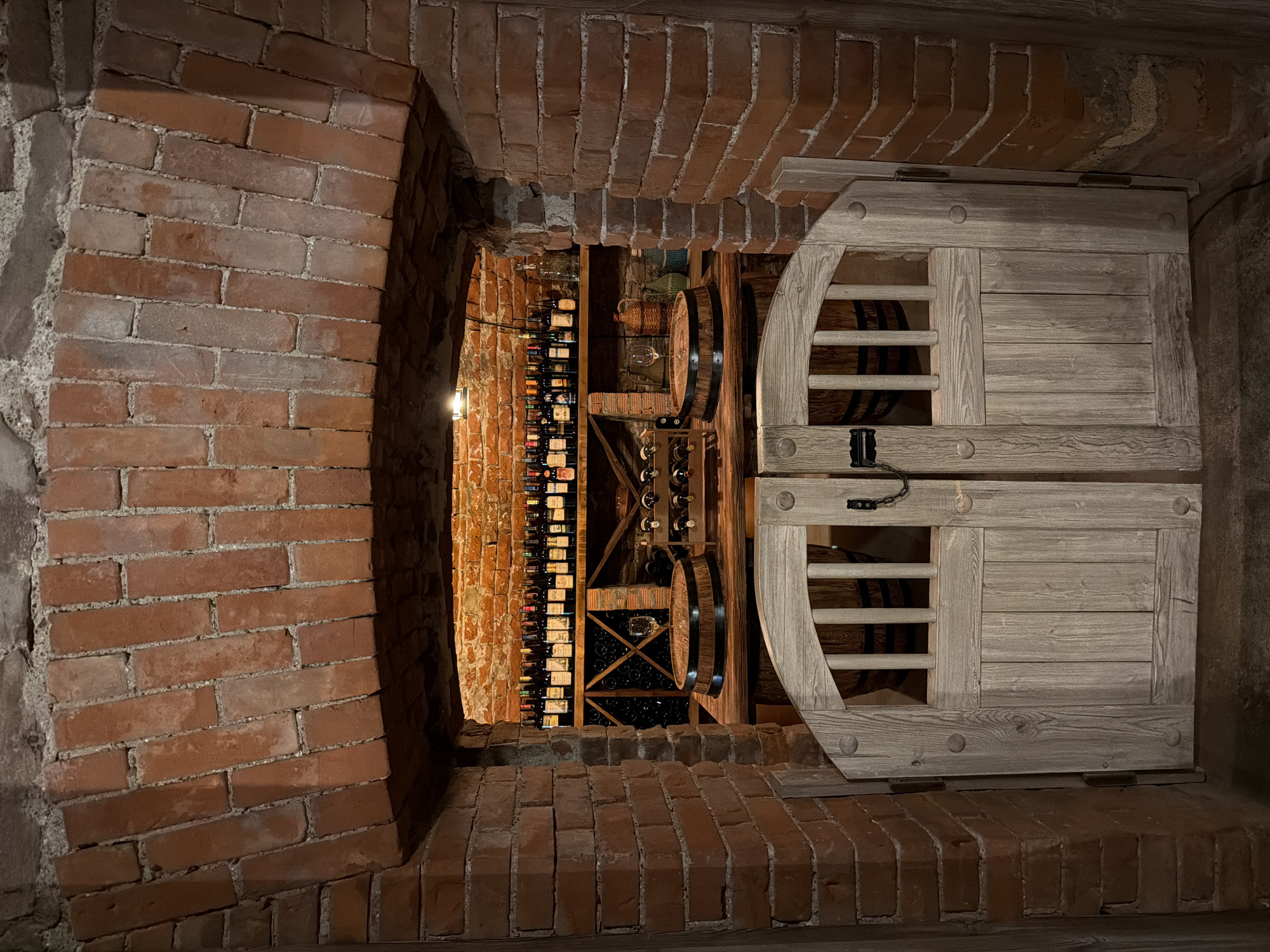 The image depicts the entrance to a wine cellar, constructed with red brick. An arched brick opening reveals a well-lit interior showcasing rows of wine bottles on shelves and in a wine rack. In the foreground, a pair of rustic wooden double doors, partially ajar, are secured by a chain. Visible through the doors are wooden barrels and decorative elements. The scene suggests a storage space designed for wine, with a rustic and traditional aesthetic. The image is not tagged with a specific location in Krasnoyarsk, Russia.