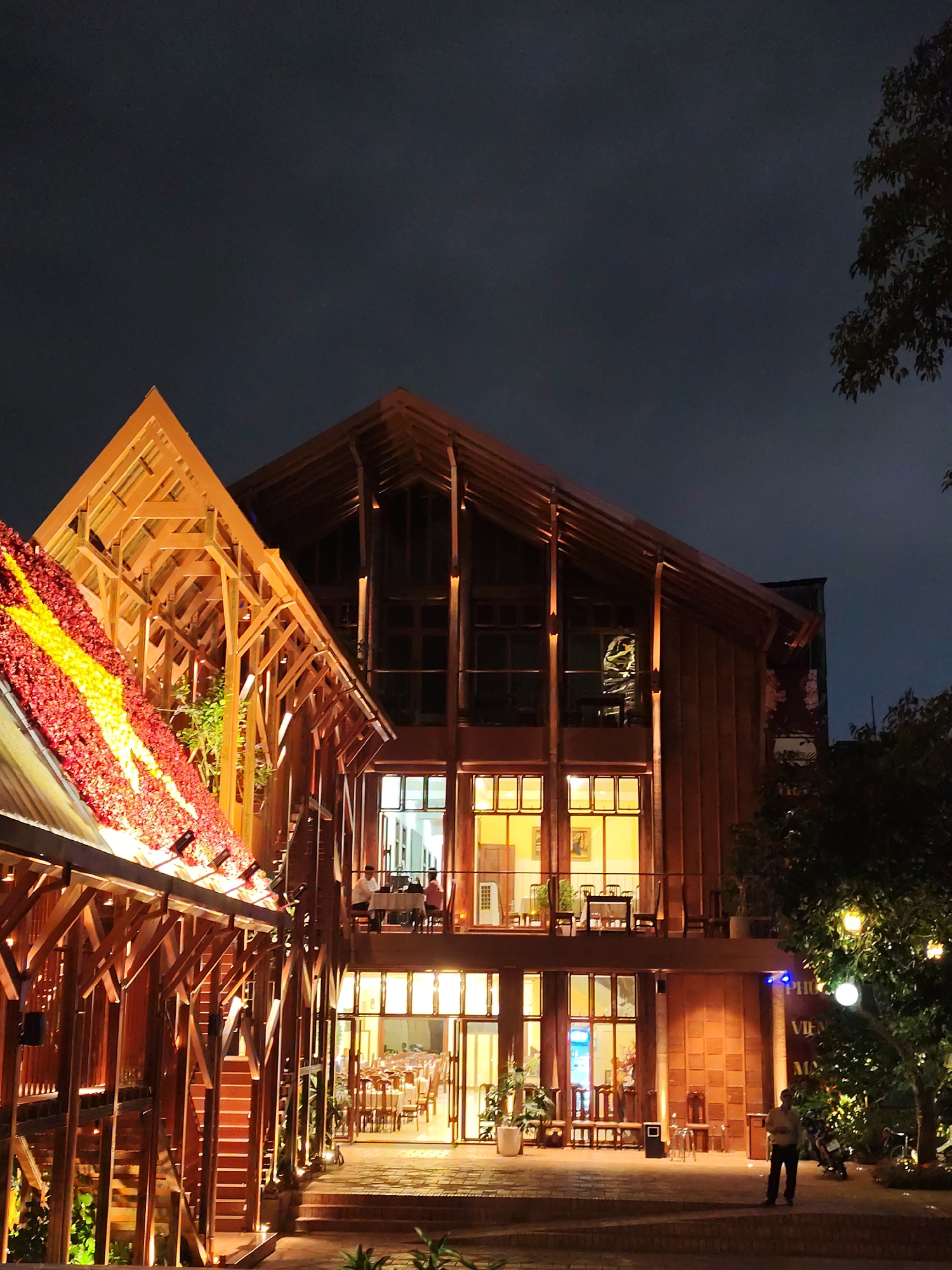 The image shows a restaurant in Da Lat, Vietnam, at night. The building has a distinctive wooden frame structure, with two gabled roofs. The roof on the left has a stripe of red and yellow flowers. The building on the right features large windows showing multiple floors of seating. A man is standing outside, while others can be seen inside dining. The sky is dark, and the building is illuminated by warm lighting. A tree on the right is also lit with decorative lights.