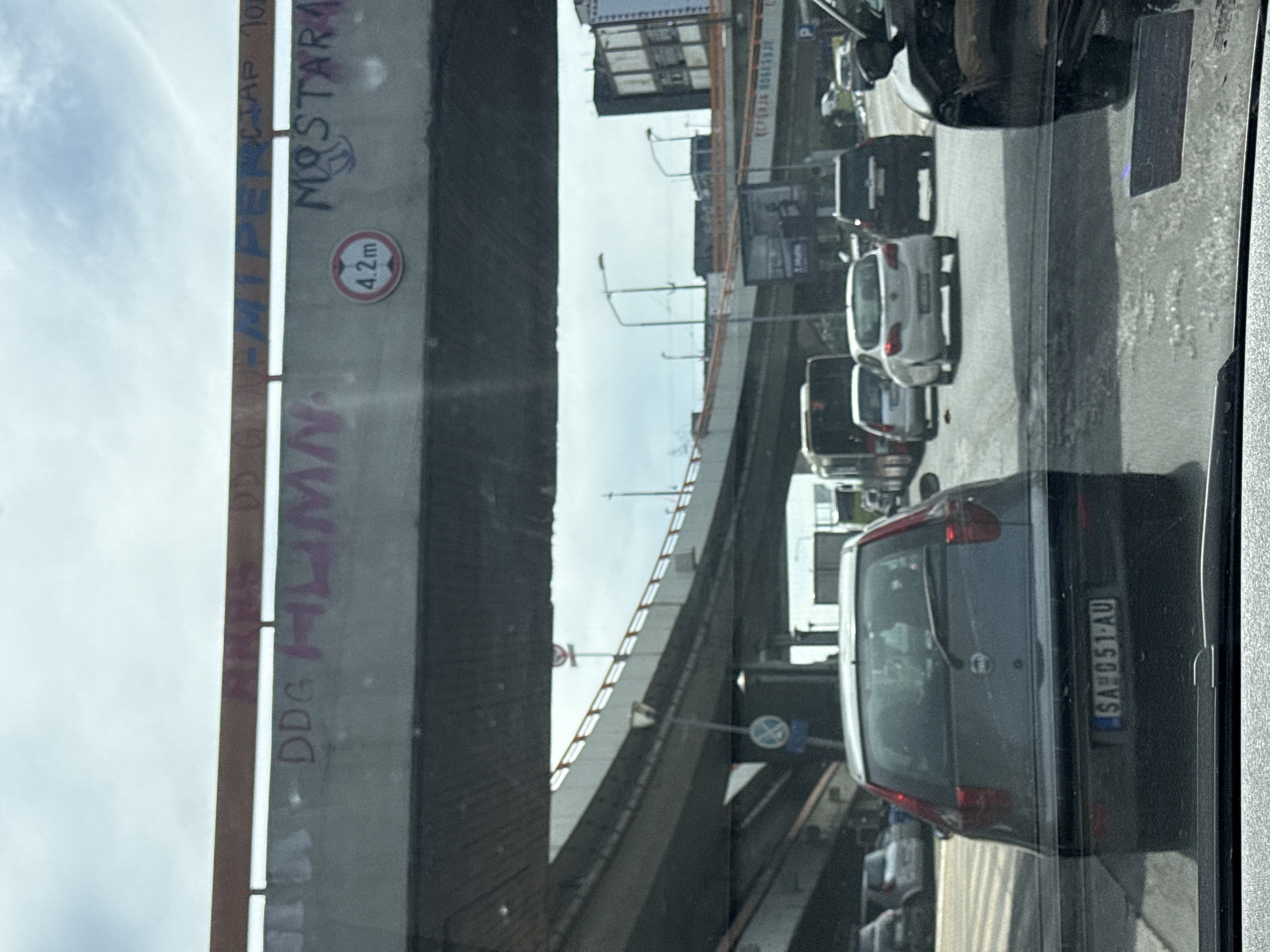 The image captures an urban street scene, viewed from a vehicle looking out through a side window, with the camera oriented sideways. An imposing concrete overpass or bridge structure spans across the upper and left parts of the frame, with the sky visible above it.
The road beneath the overpass is busy with traffic, consisting of several cars and what appears to be a truck, all seemingly stationary or moving slowly in a traffic jam. In the immediate foreground, the rear of a dark hatchback or SUV is clearly visible, featuring a license plate that reads "SA 051-AU". Further ahead, a white car and other vehicles are present.
The concrete structure of the overpass displays graffiti, including the word "MOSTAR" and other markings such as "DDG" and "HUMM". A circular traffic sign indicating a "4.2m" height restriction is also affixed to the overpass support. In the background, beyond the traffic, more elevated road structures, streetlights, and some advertising billboards can be seen.
The scene depicts a clear, possibly slightly overcast, daytime environment in a city. The visible license plate (SA typically refers to Sarajevo) and the graffiti "MOSTAR" suggest a location within Bosnia and Herzegovina.