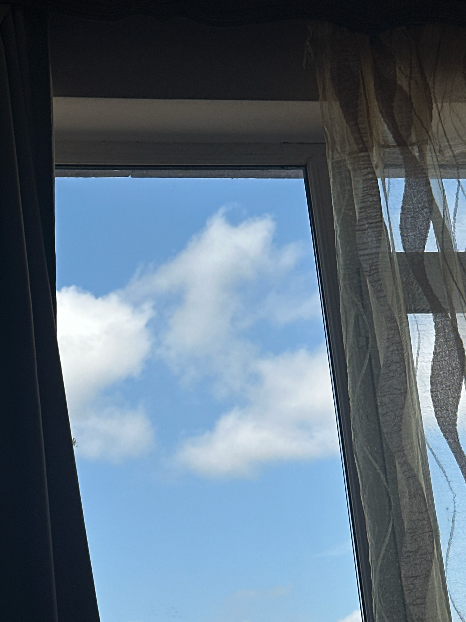 The image shows a view through a window, looking up at a bright blue sky with scattered white cumulus clouds. A dark curtain is partially drawn on the left side of the frame, and a sheer, patterned curtain is visible on the right side. The window frame appears to be made of a light-colored material, possibly white plastic or painted wood. The perspective suggests the photo was taken from inside a building, looking outwards. There are no people or animals visible in the image, and no discernible architectural features of the city of Gyumri, Armenia, are present. The scene is an ordinary outdoor view from an interior setting.