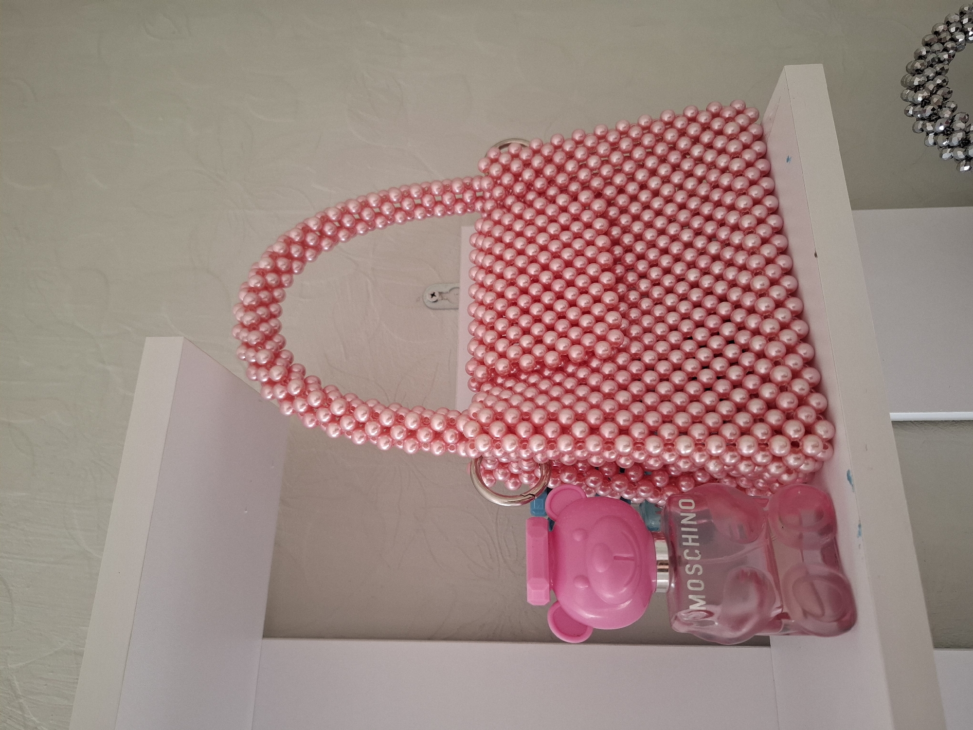 A pink Moschino perfume bottle shaped like a bear and a pink pearl beaded handbag are displayed on a white shelf. The background is a textured, light-colored wall.