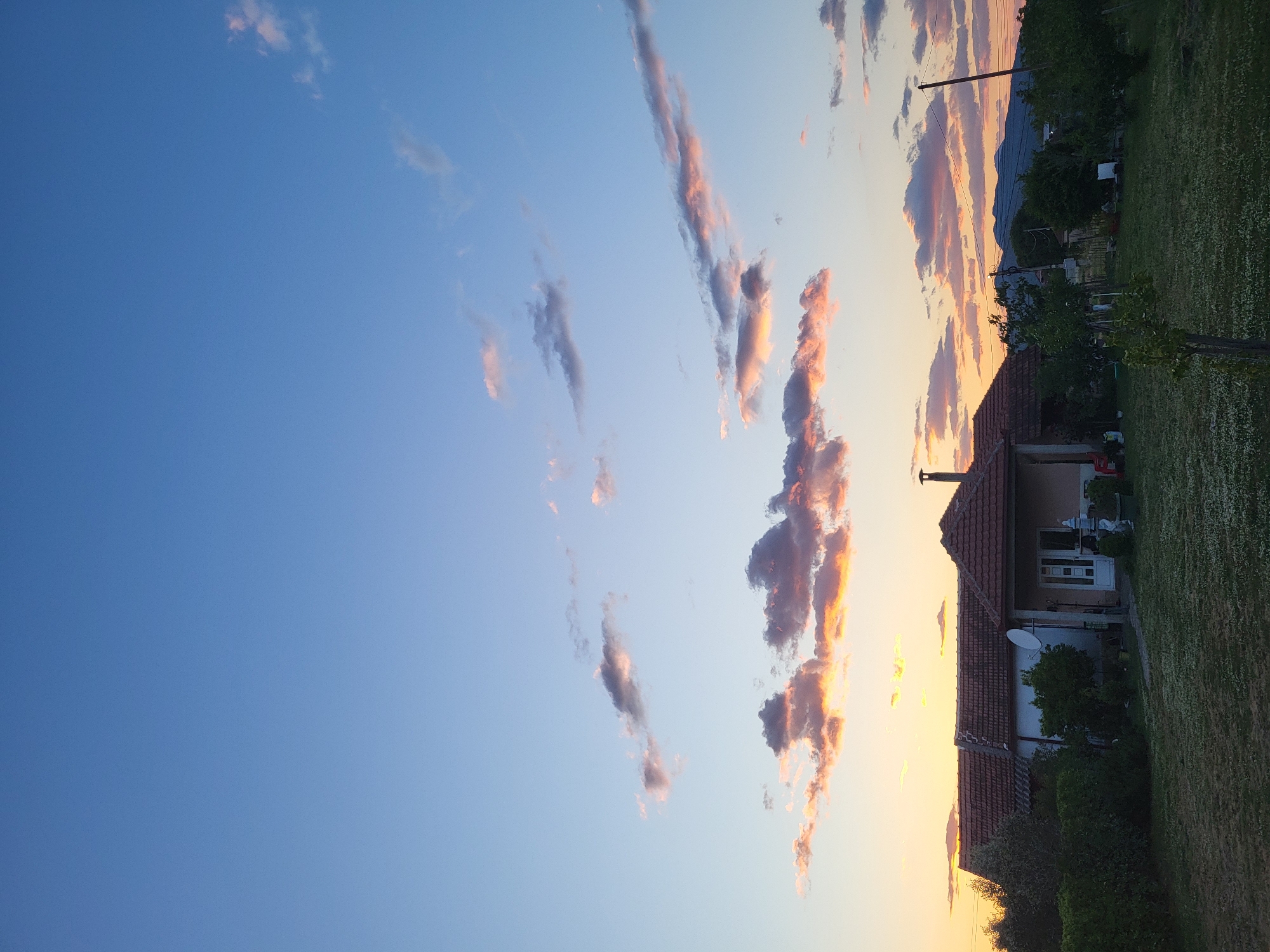 The image captures a horizontal landscape view in Mahala, Montenegro at dusk. The sky transitions from a deep blue on the left to a light orange-yellow near the horizon on the right. Streaks of cumulus clouds, tinged with pink and purple hues, are scattered across the sky.

On the right, a single-story house with a terracotta-tiled roof is partially visible. A satellite dish is mounted on its side. Surrounding the house is a lawn that extends into the foreground, showing a mix of green grass and clover.

In the background, above the house, a mountain range and trees are silhouetted against the sunset. A power line is stretched across the landscape, held aloft by a utility pole. No people are visible in the frame.